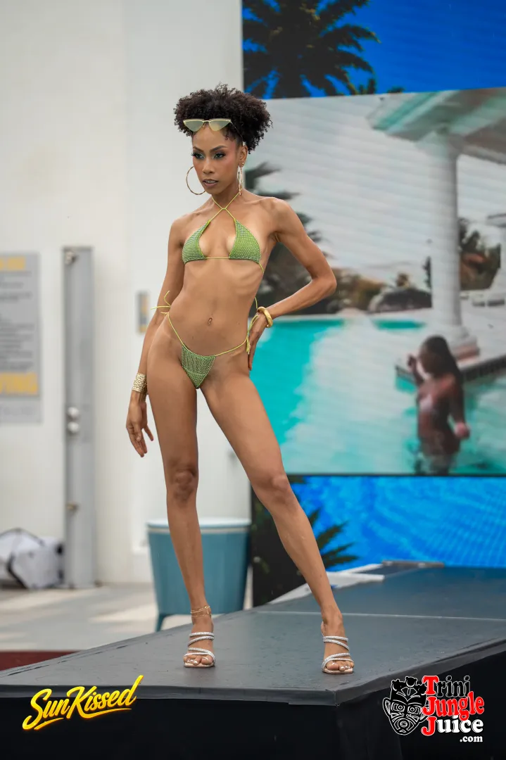 Woman in green bikini walking on runway with pool background, palm trees, and audience, during a fashion show.