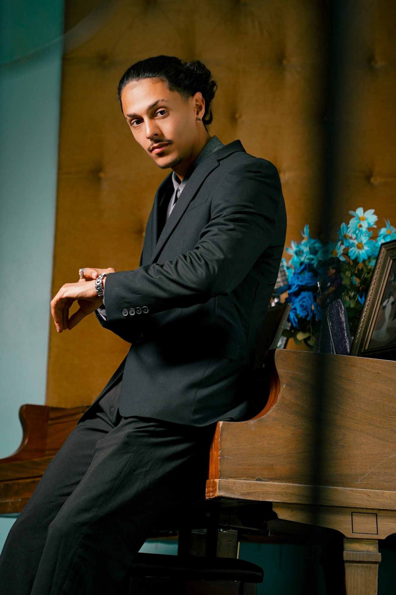Person with dark hair tied back, wearing a dark suit, sitting on a wooden chair in a room with a yellow wall and blue flowers.