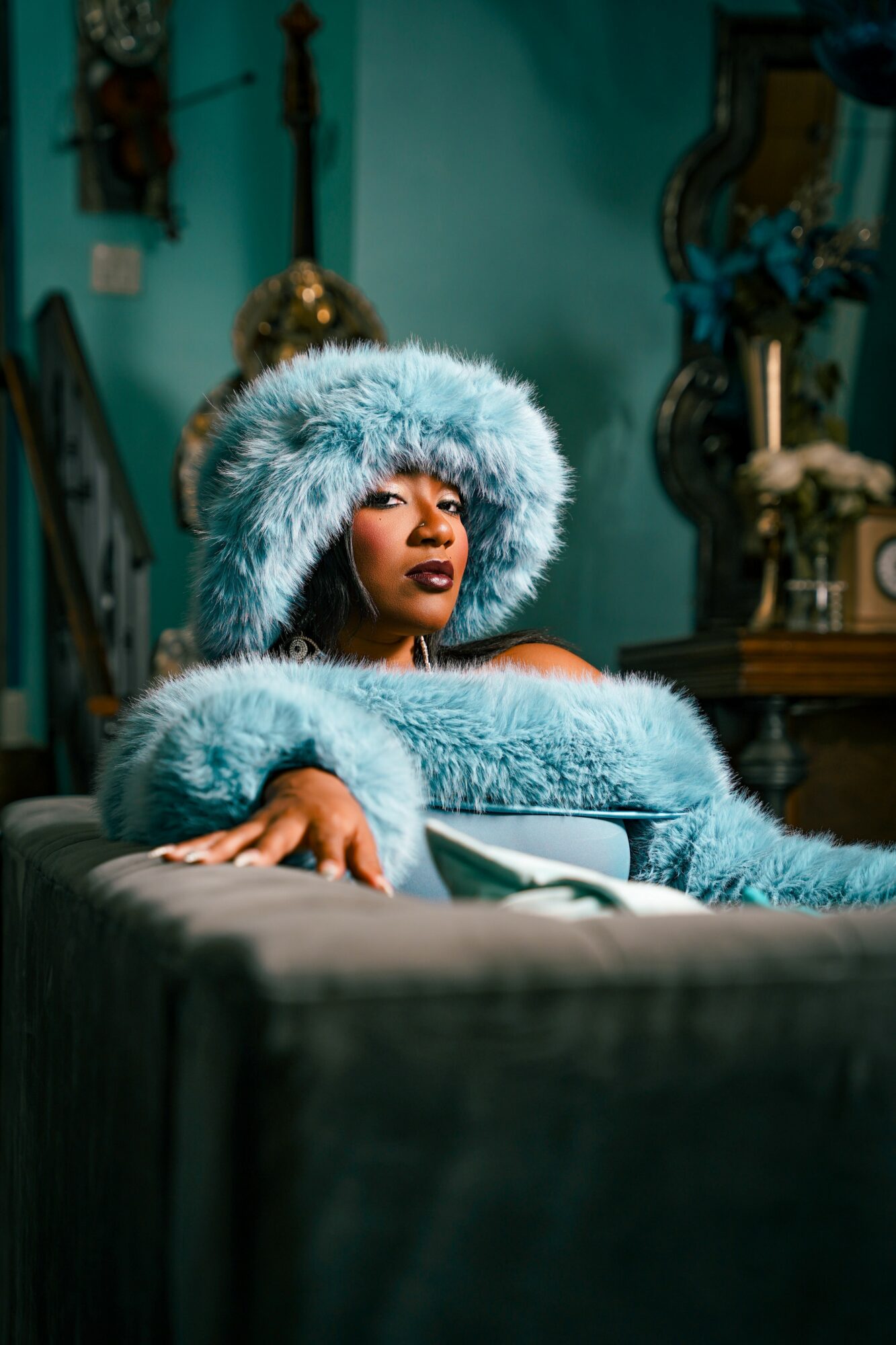 Woman wearing a large, fluffy blue hat and matching coat, sitting on a sofa in a decorated room.
