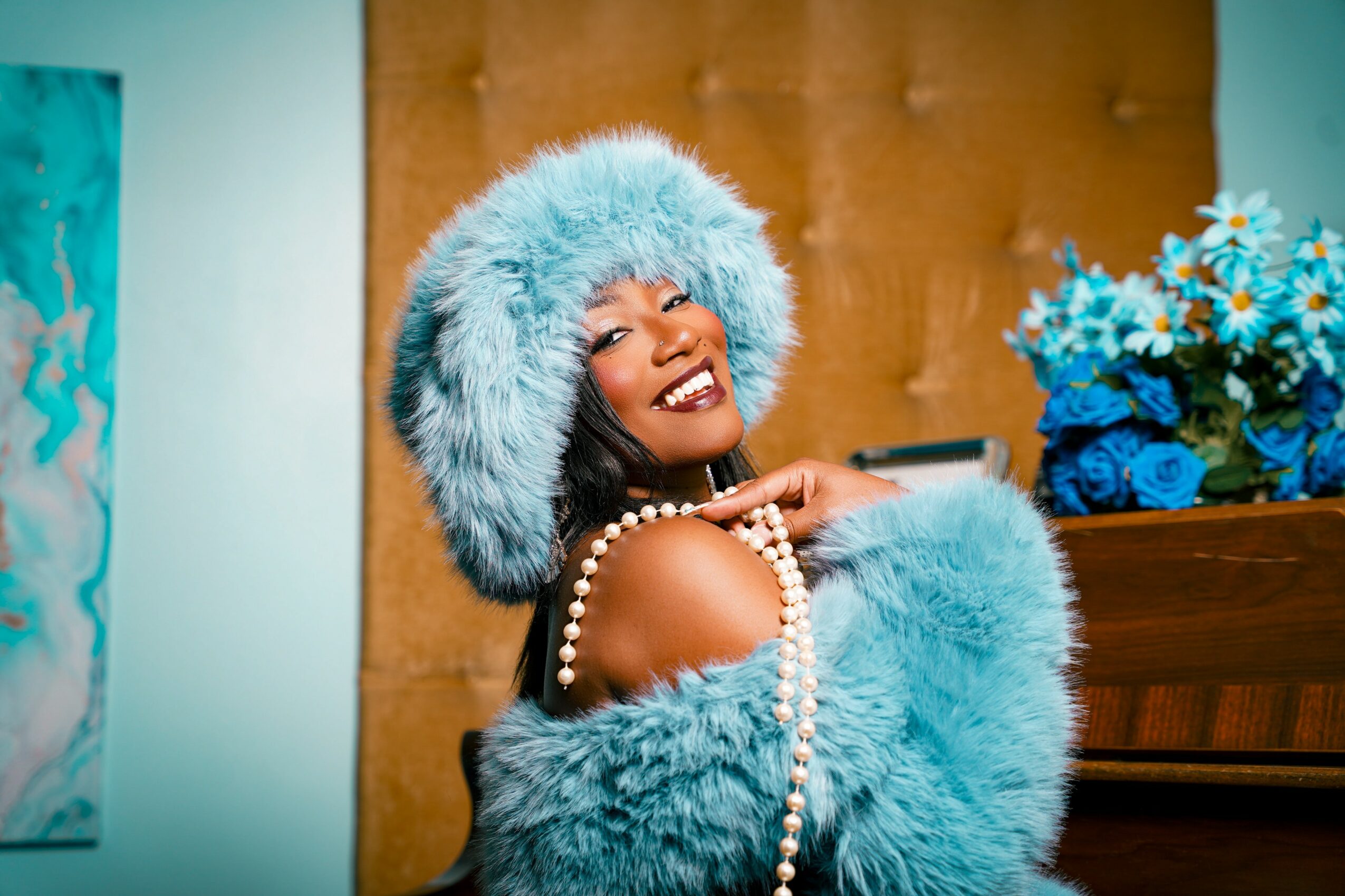 Smiling woman wearing a large blue furry hat and matching coat, holding a pearl necklace, with a bouquet of blue flowers nearby