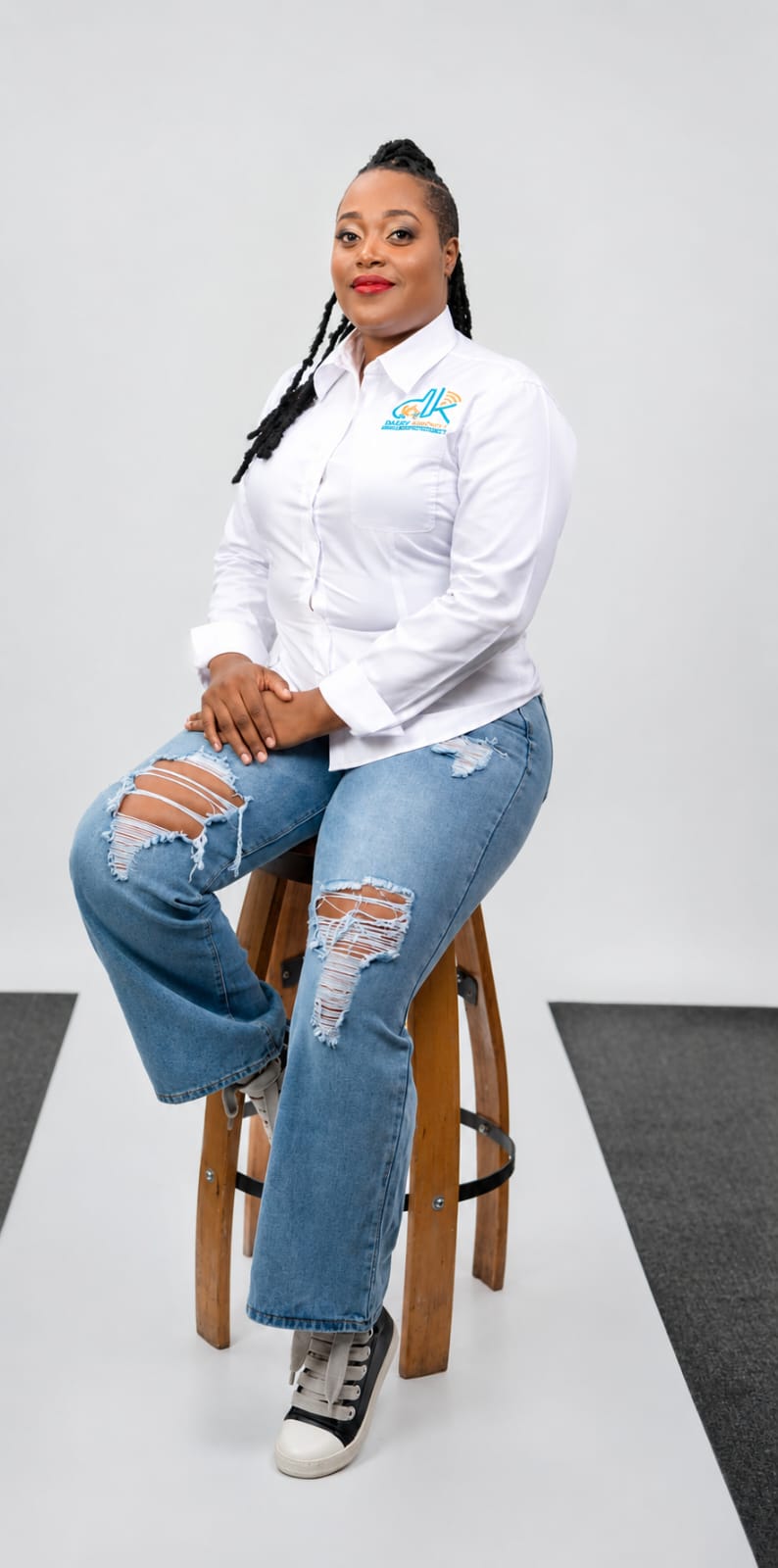 Woman sitting on a stool, wearing a white shirt and ripped jeans, smiling, with a plain background.