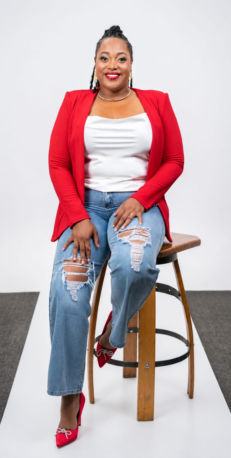 Woman sitting on a stool, wearing a red blazer, white top, ripped jeans, and pink shoes, smiling at the camera.