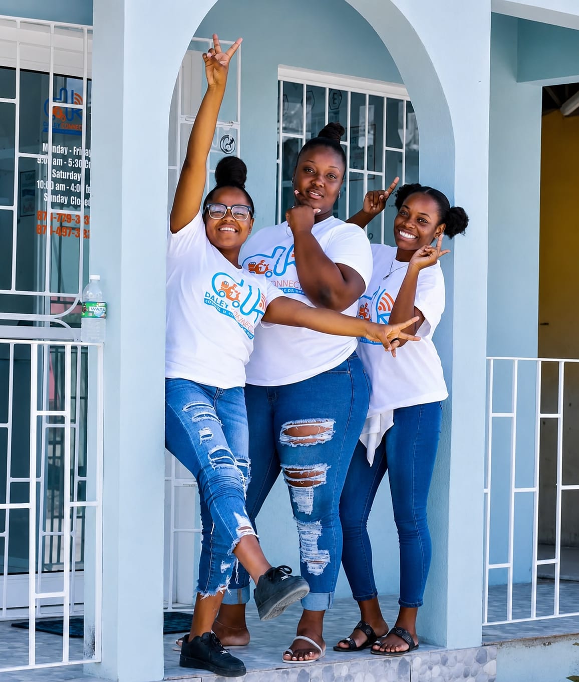 Three women standing on a porch, smiling and posing with arms raised and crossed, in front of a light blue wall and white railing.