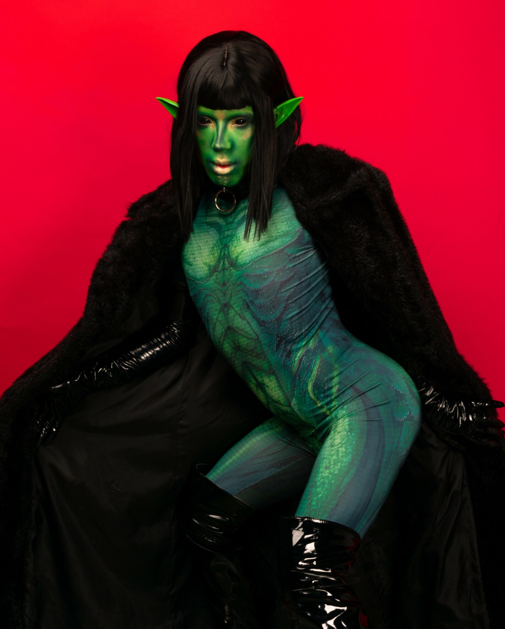 Person with green face paint, elf ears, black hair, wearing black clothing and a black cape, sitting against a red background.