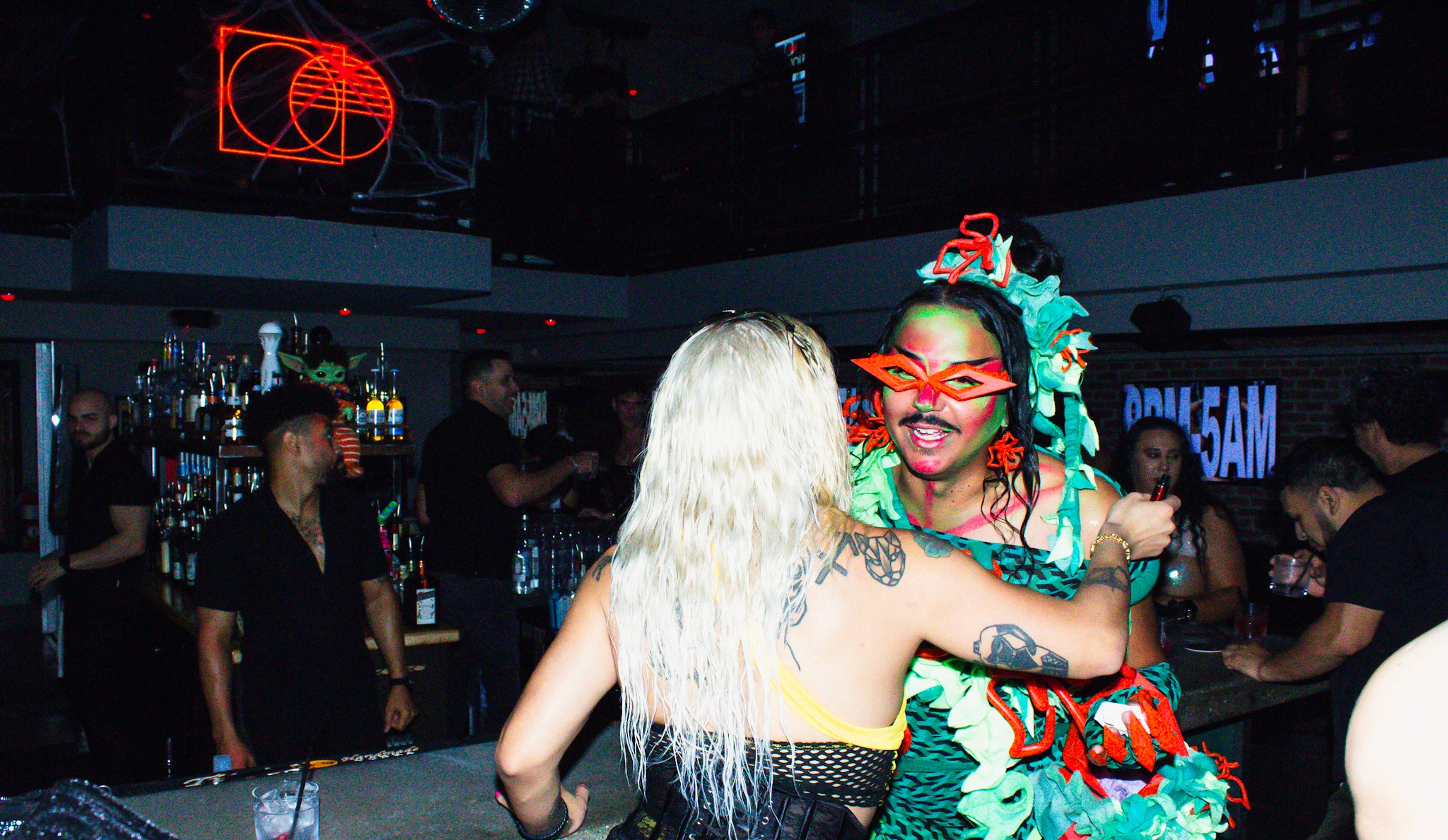 Two women embrace in a nightclub with a bar and people in the background, one with blonde hair and the other with colorful accessories.