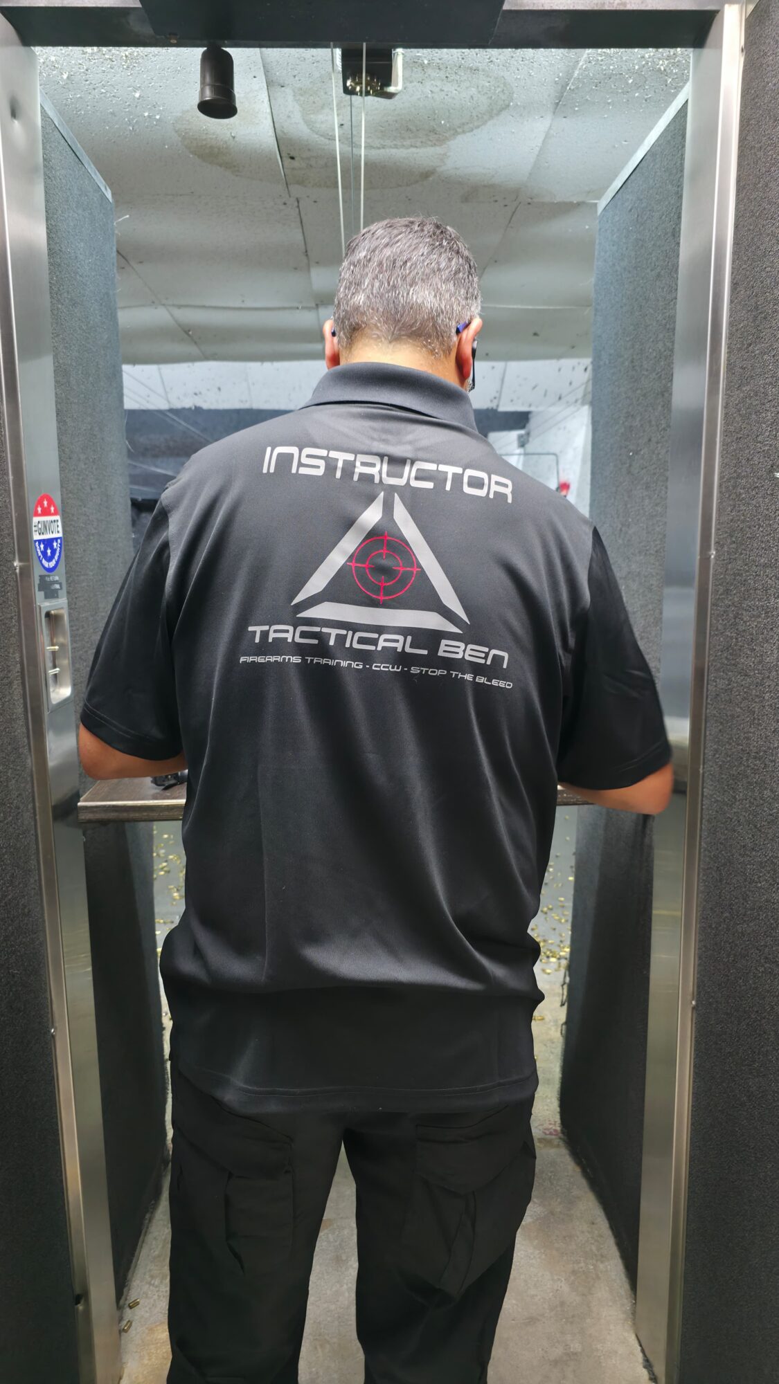 Person with short hair wearing a black tactical shirt, standing in a shooting range booth, facing away.