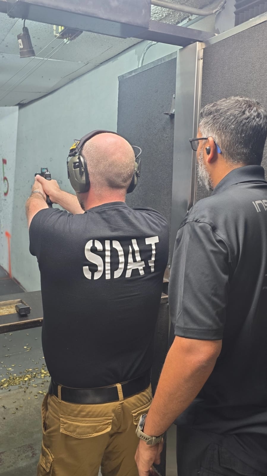 Two men at a shooting range, one aiming a handgun, the other observing, both wearing black shirts, one with 'SPAT' on back.
