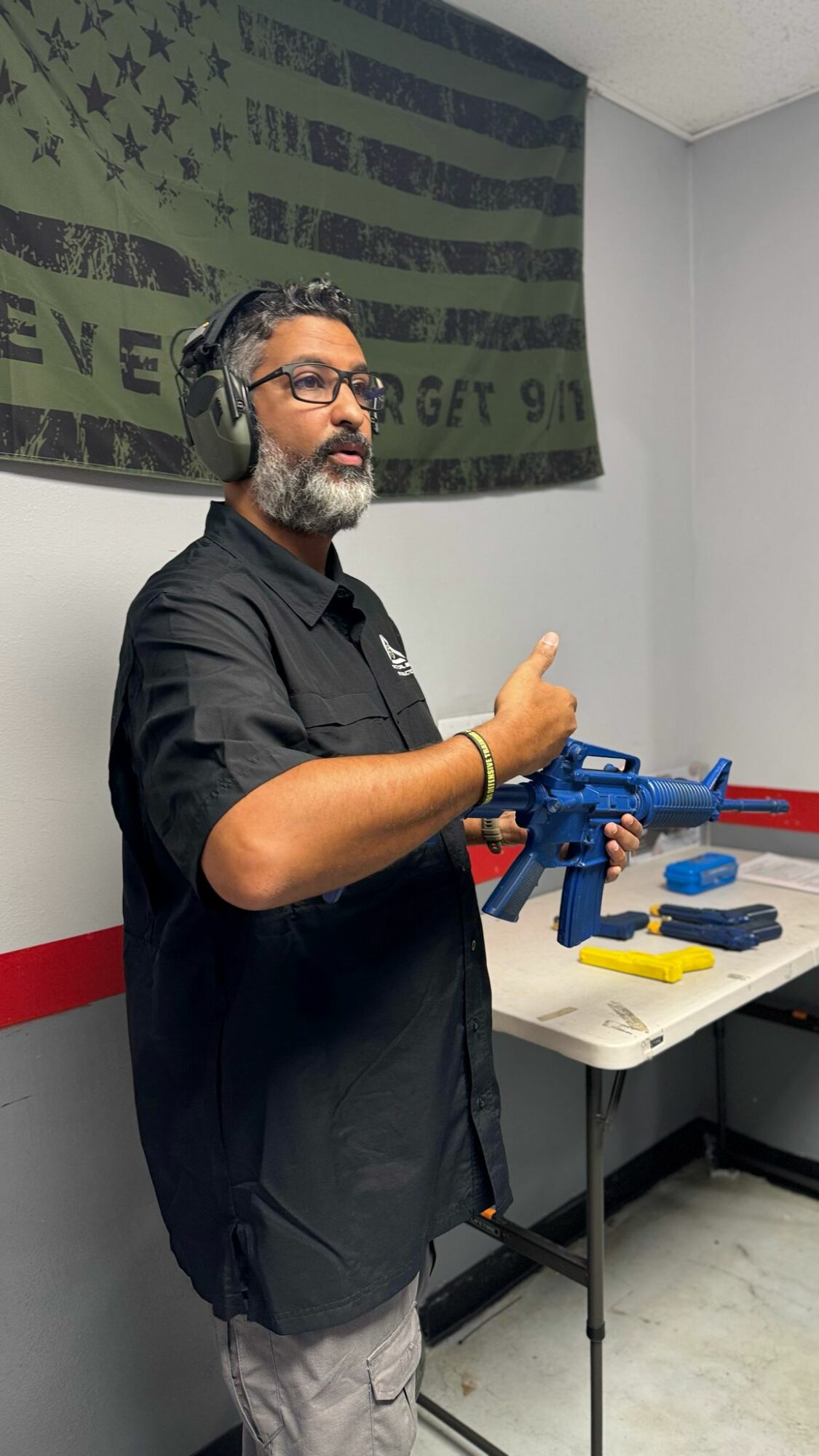 Man with glasses and headphones giving a thumbs-up, standing next to a table with a blue toy gun and accessories, in a room with a flag on the wall.
