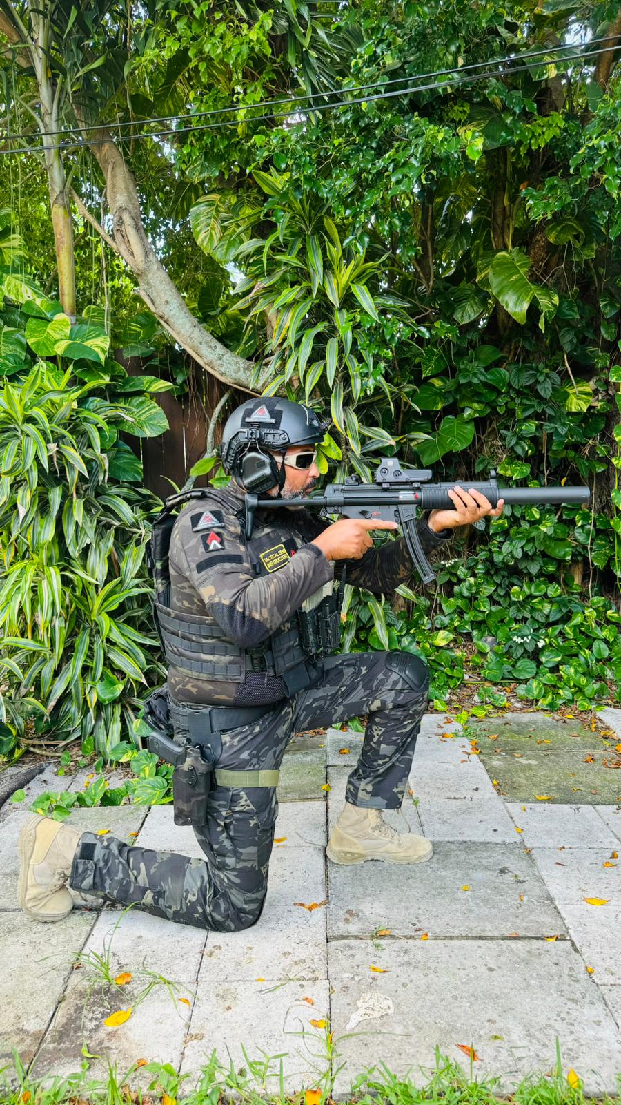 Person in tactical gear kneeling outdoors with a rifle, surrounded by green foliage and trees.