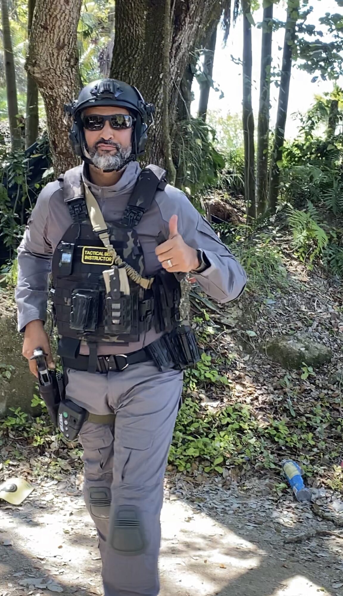 Police officer in uniform and helmet giving a thumbs-up outdoors with trees and sunlight, holding a firearm, smiling.