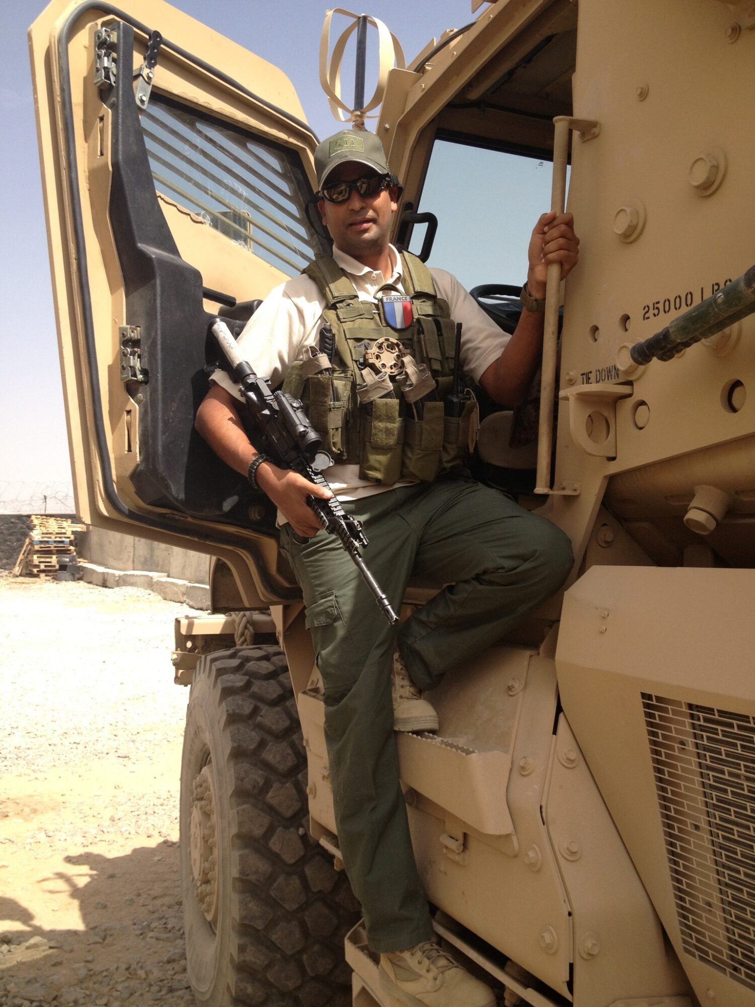 Person in military gear sitting on a vehicle, holding a rifle, wearing sunglasses and a cap, in a desert environment.