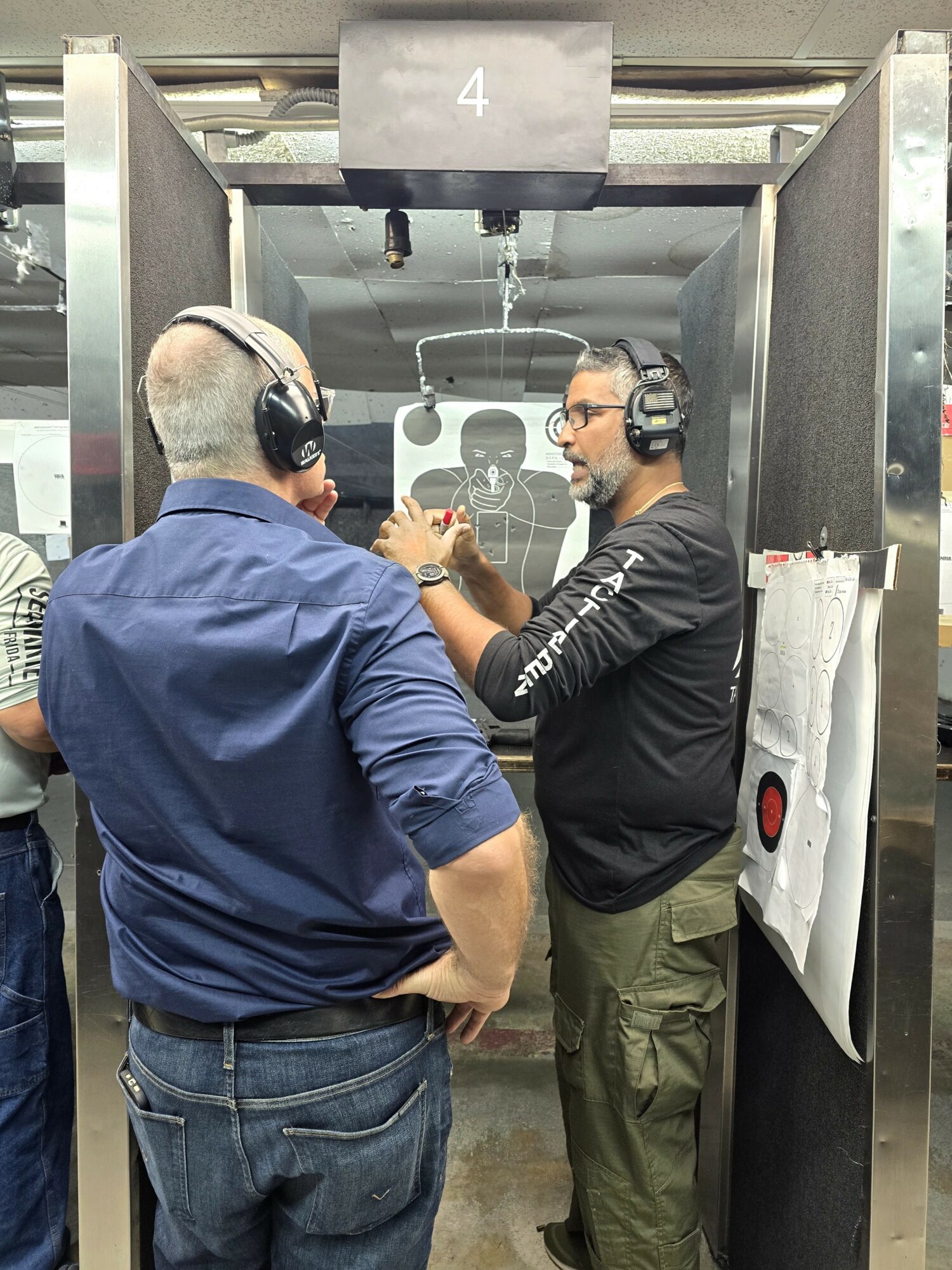 Two men wearing headphones and glasses are at a shooting range, aiming at a target through a window.