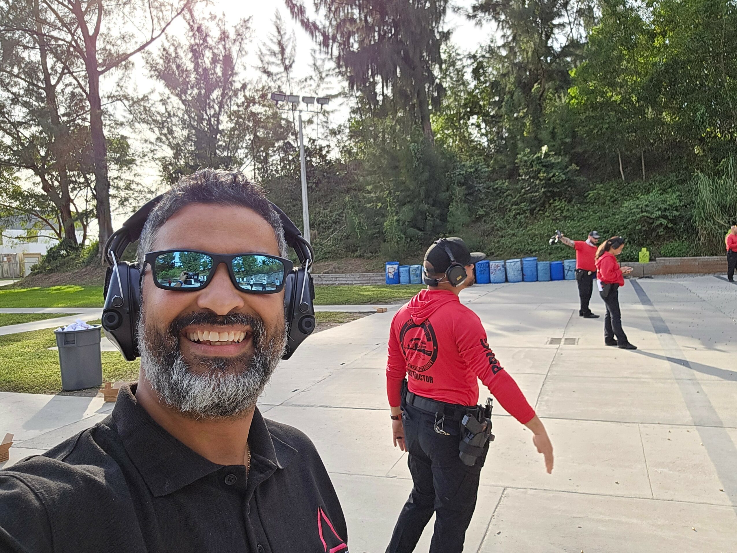 Smiling man with headphones and sunglasses taking a selfie outdoors, with people in red shirts in the background.