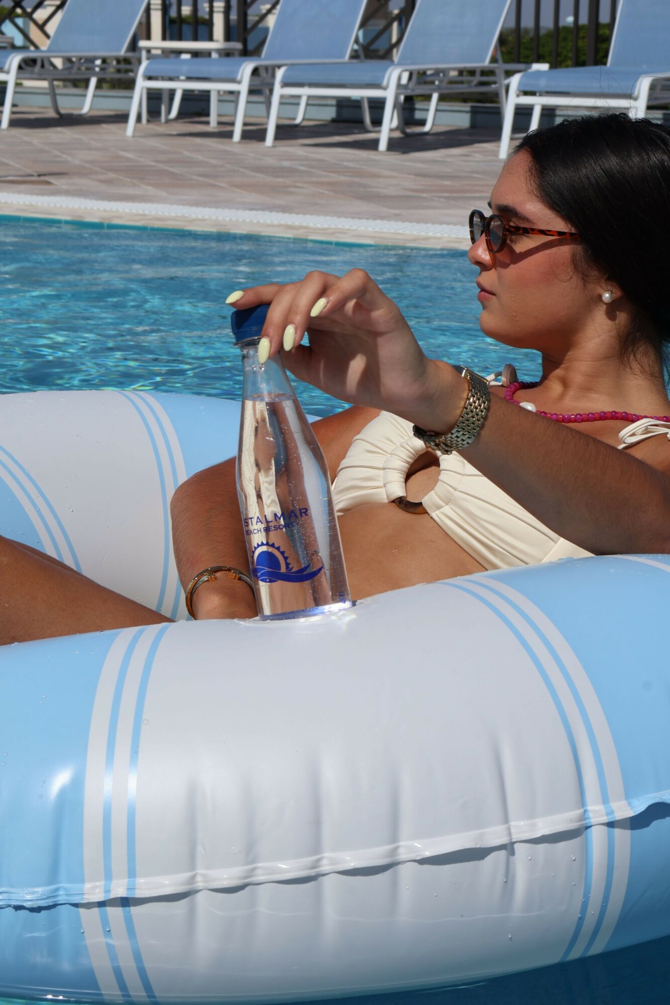 Person relaxing by poolside, holding a drink, wearing sunglasses and a watch, with pool and lounge chairs in background.