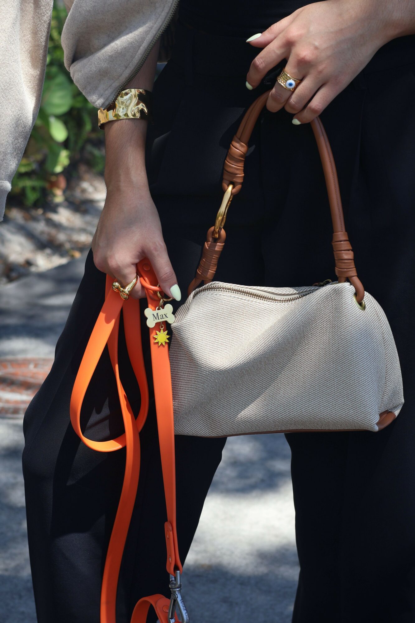 Person holding a beige purse with a wooden strap, wearing a watch and a ring, outdoors with sunlight and shadows.