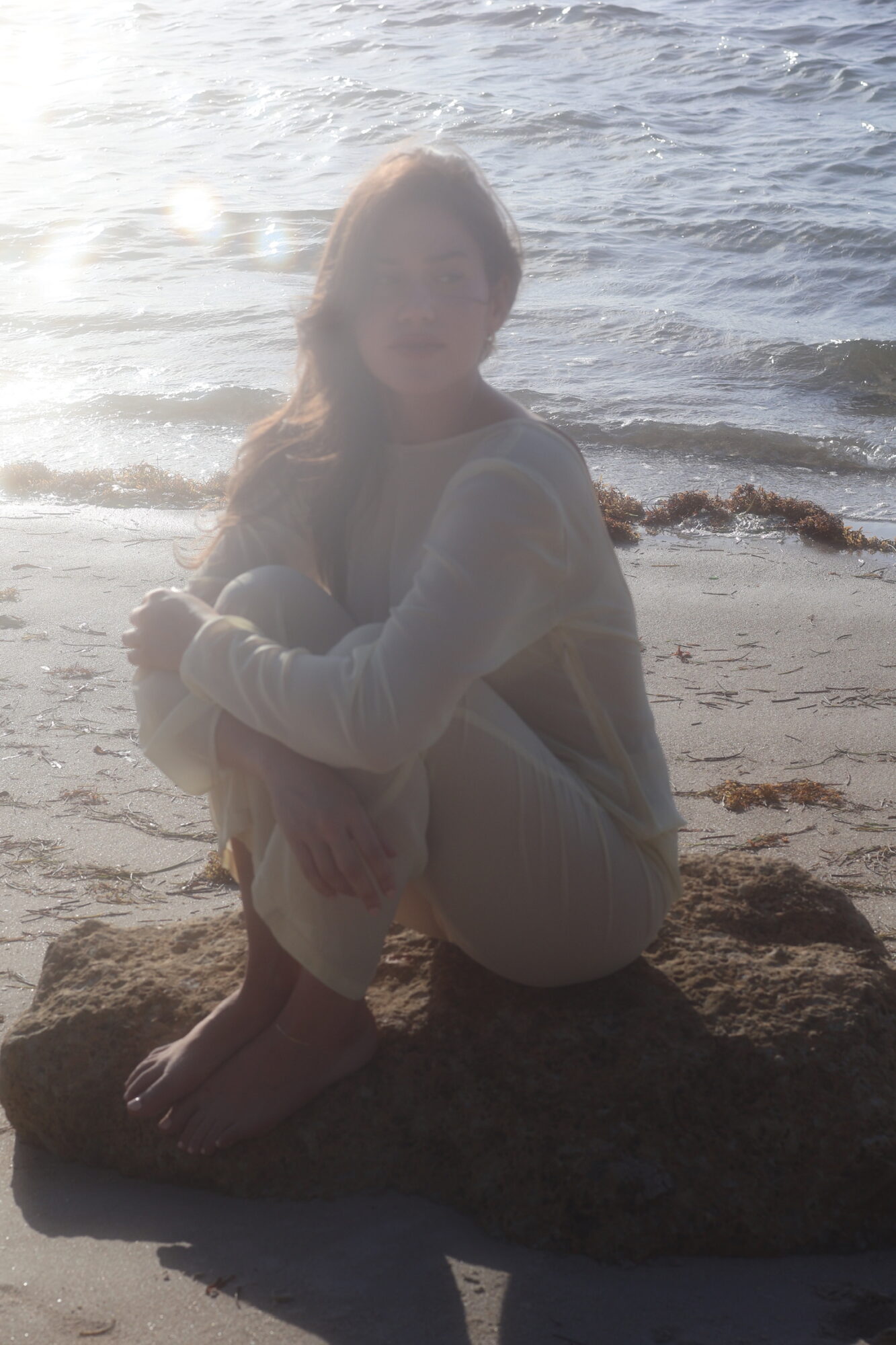 Person sitting on a rock at the beach with water in the background, sunlight shining, wearing light-colored clothing.