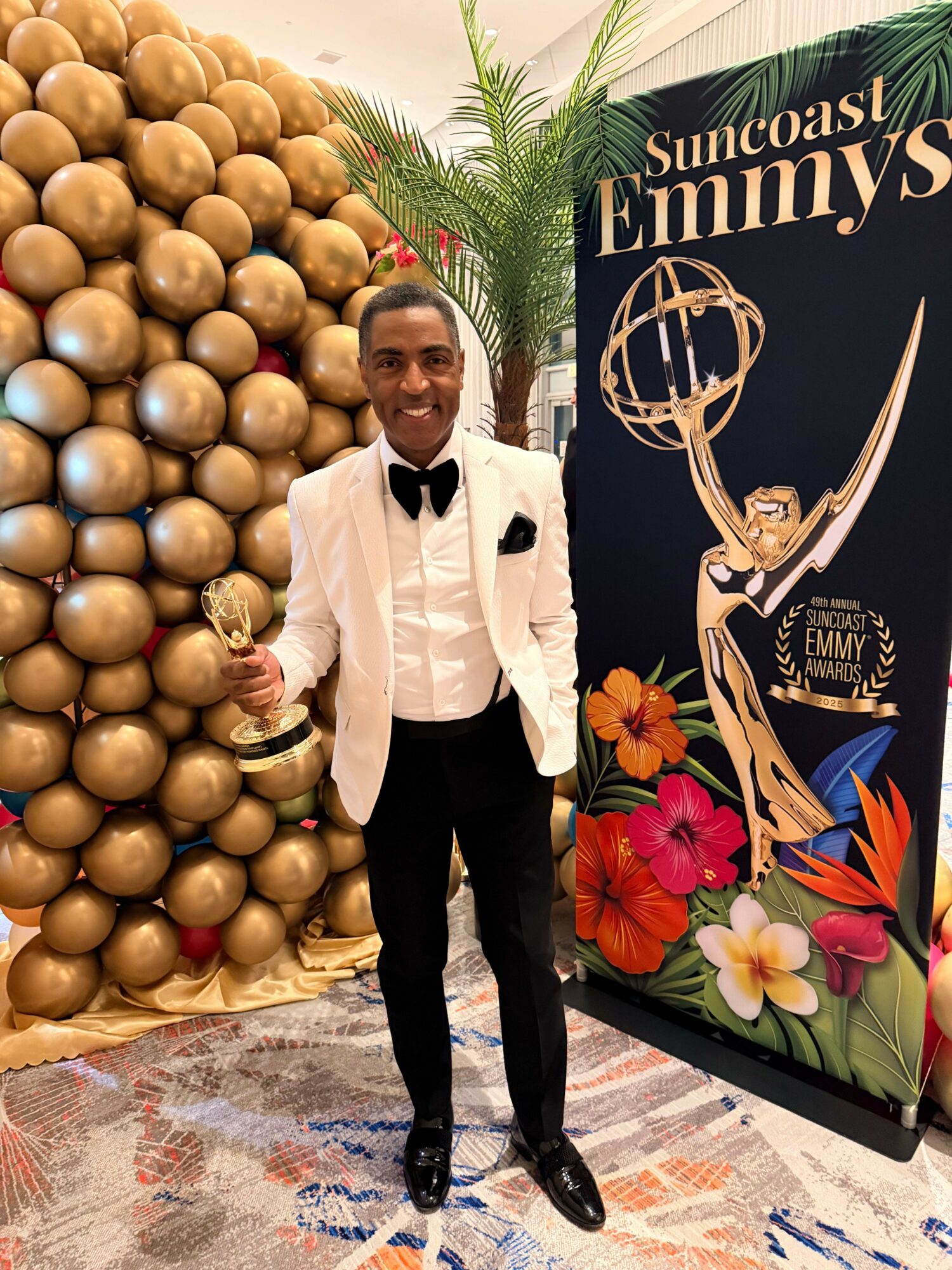 Person in white shirt and black pants standing in front of a gold ball backdrop and a colorful poster, holding a drink.