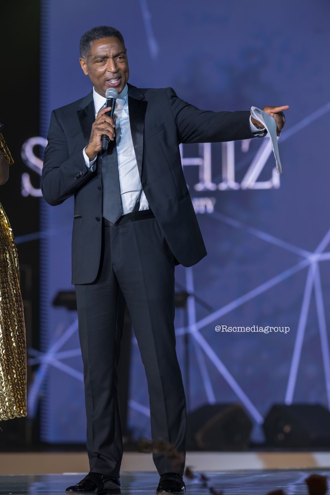 Man in suit speaking into microphone and pointing, on stage with a blue background and geometric design.