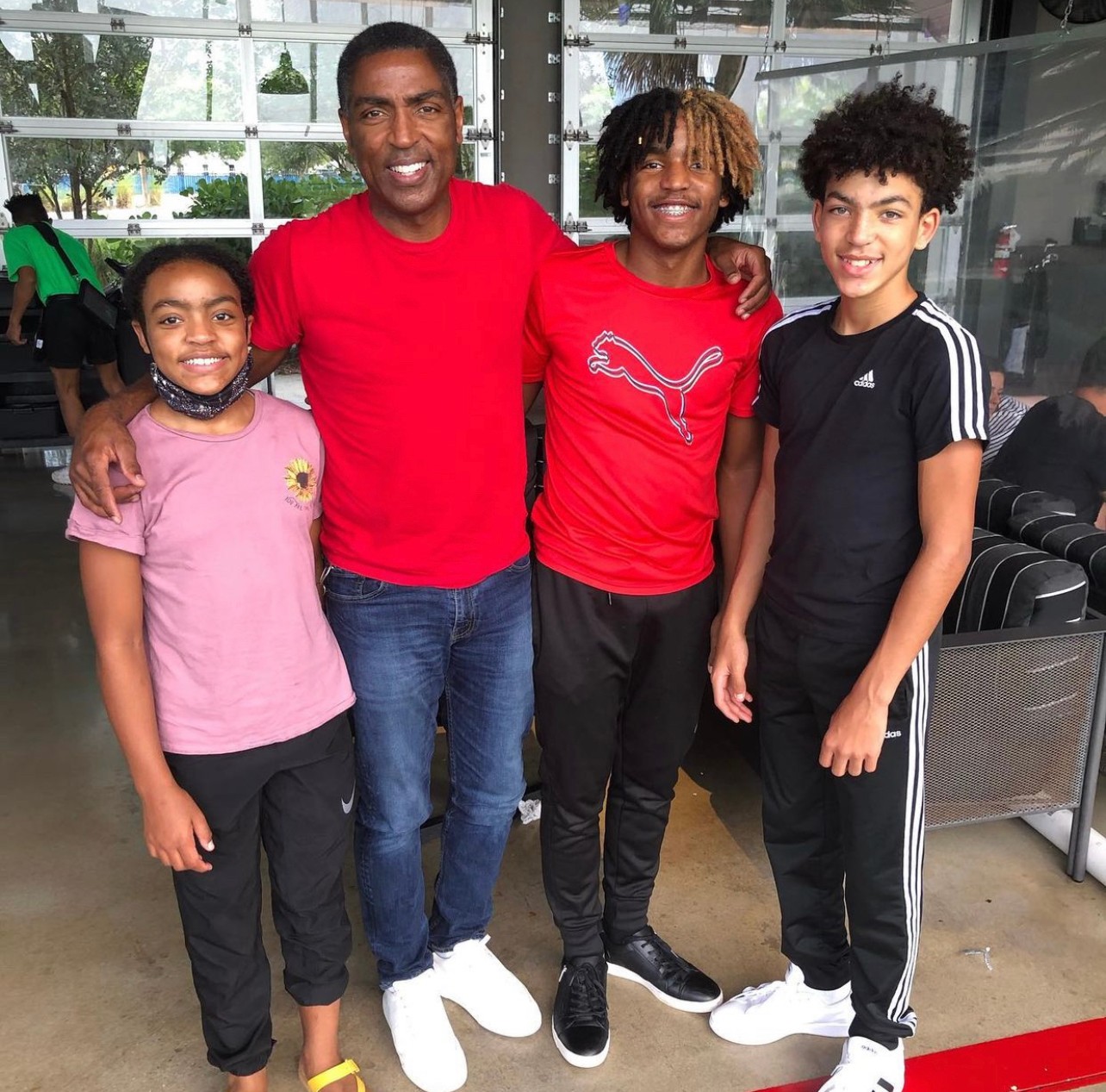 Four people standing together indoors, smiling, with large windows behind them, three children and one adult, casual clothing, bright environment.