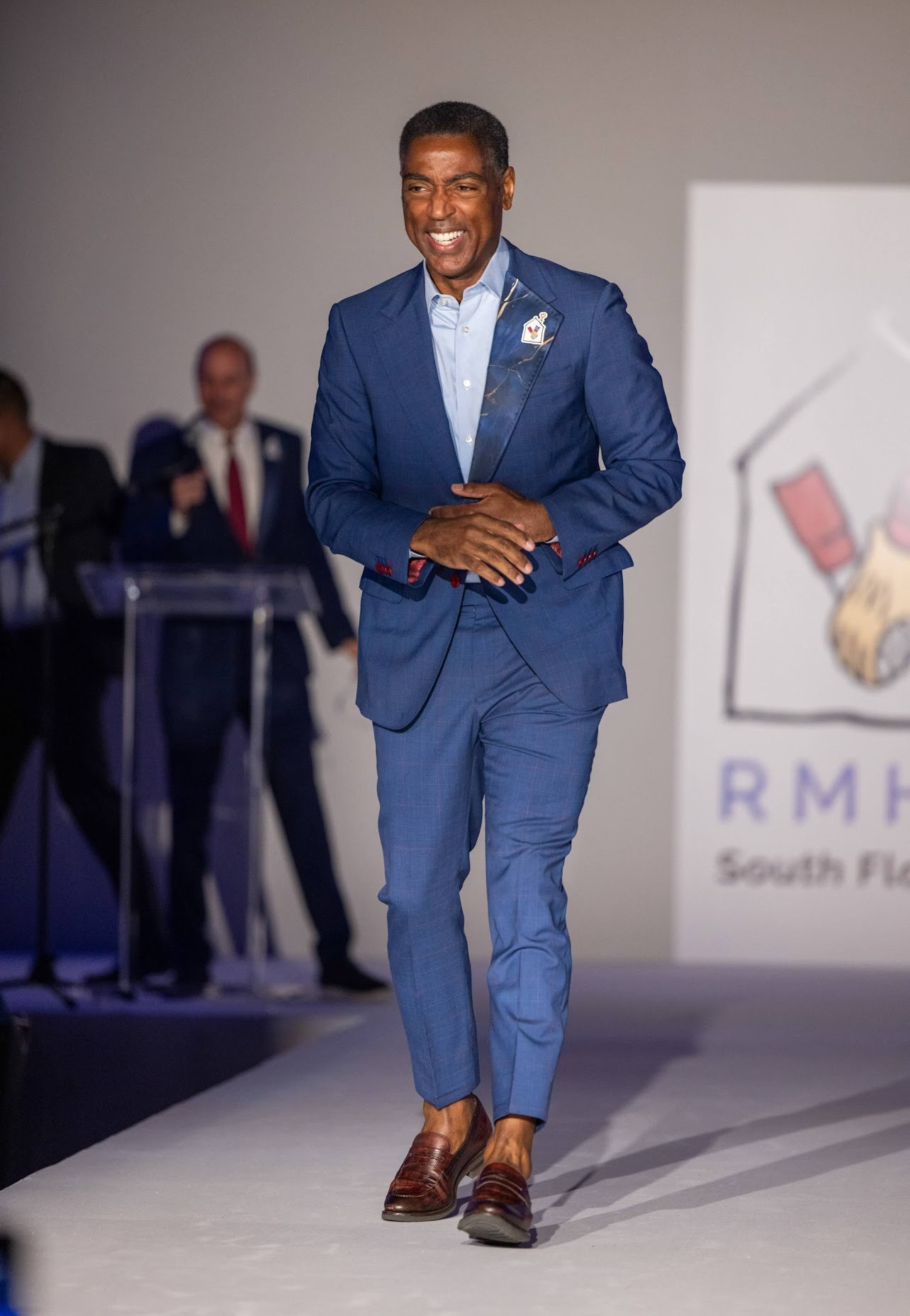 Man in blue suit walking on stage, smiling, with people and a logo in the background.