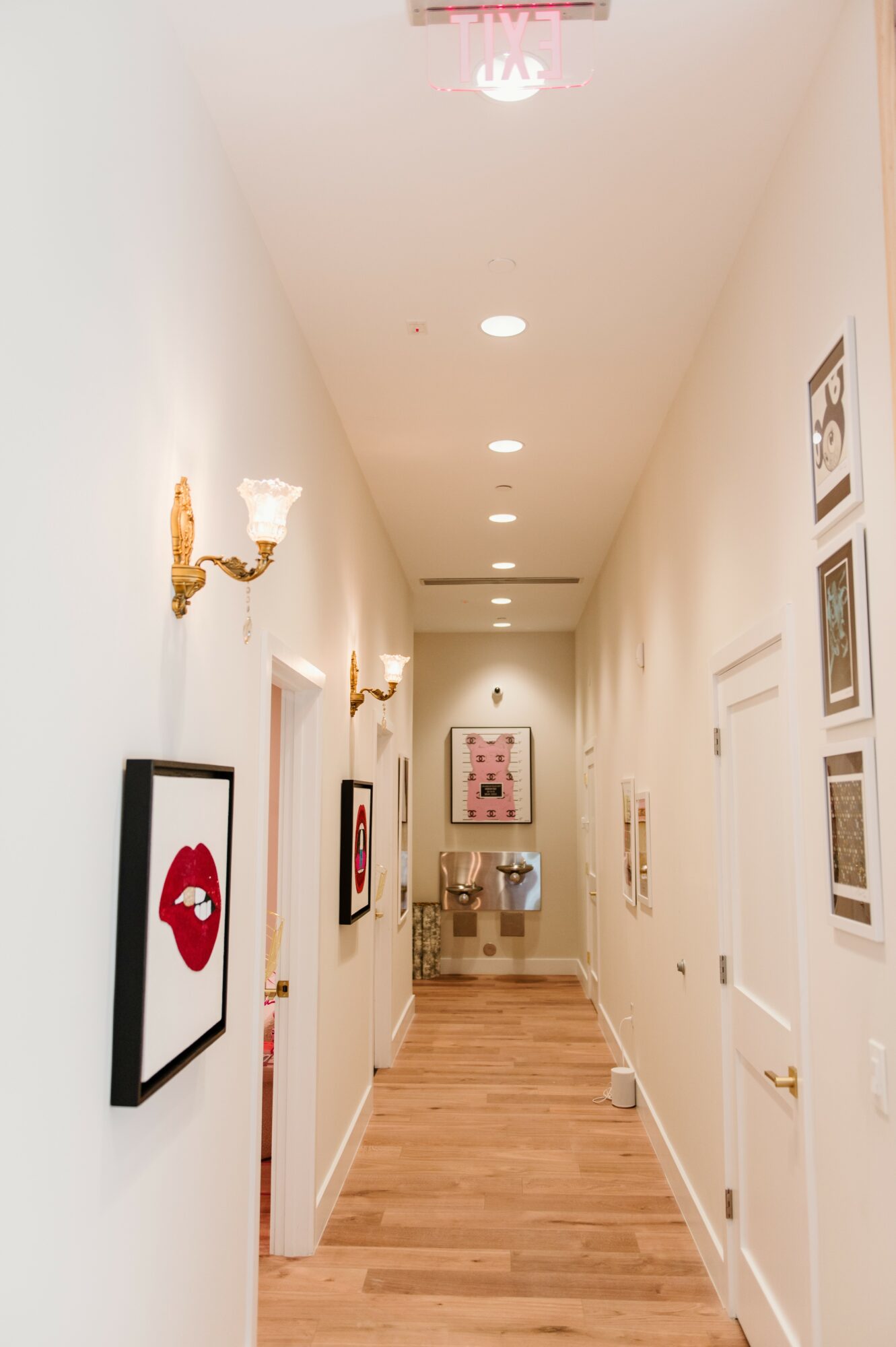 Long hallway with framed artwork on white walls, wooden floor, and ceiling lights, leading to a room with a table and chairs.
