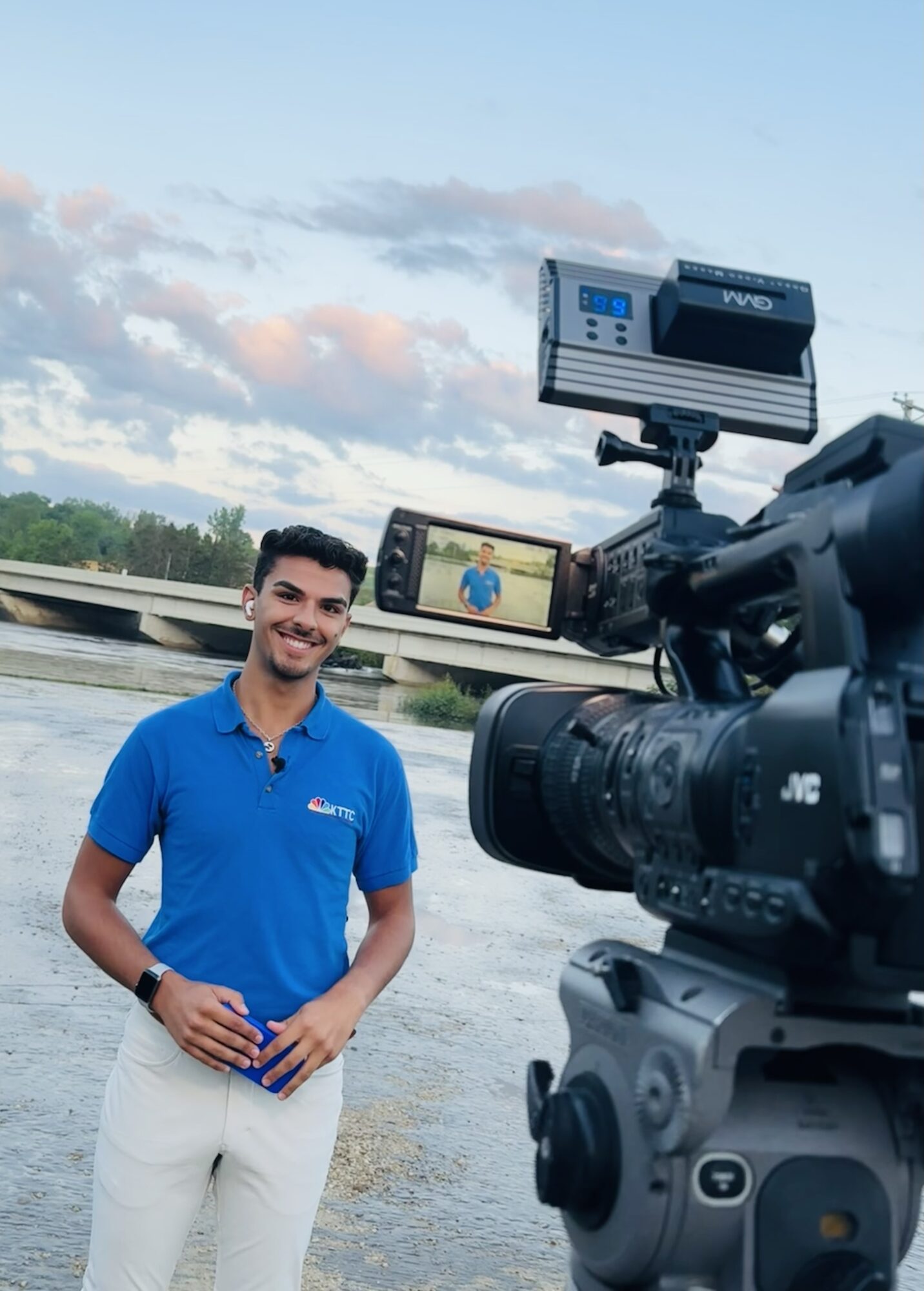 Man in blue shirt standing outdoors near water, smiling, with camera equipment filming him, sky with clouds in background.