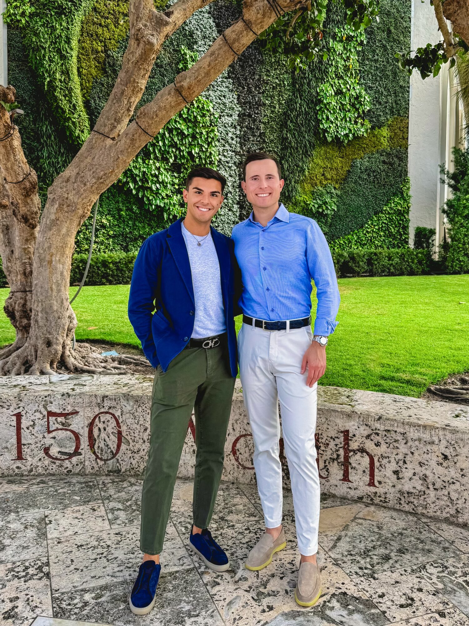 Two men stand outdoors in front of a green hedge and tree, smiling, dressed in smart casual clothing.