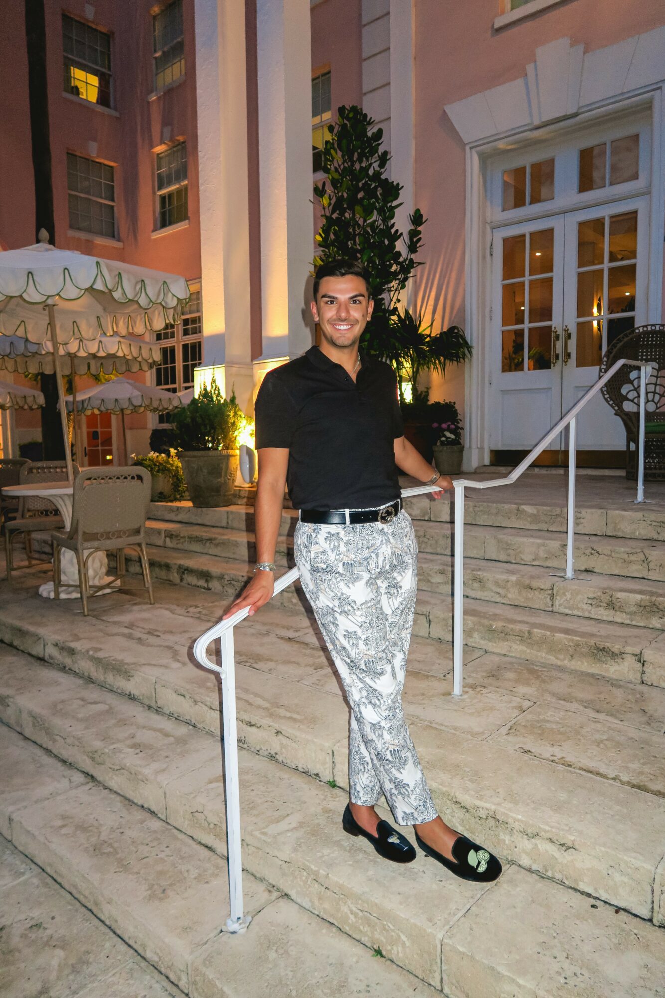 Person standing on stairs outside building at night, smiling, wearing black top and patterned pants, with outdoor seating nearby.
