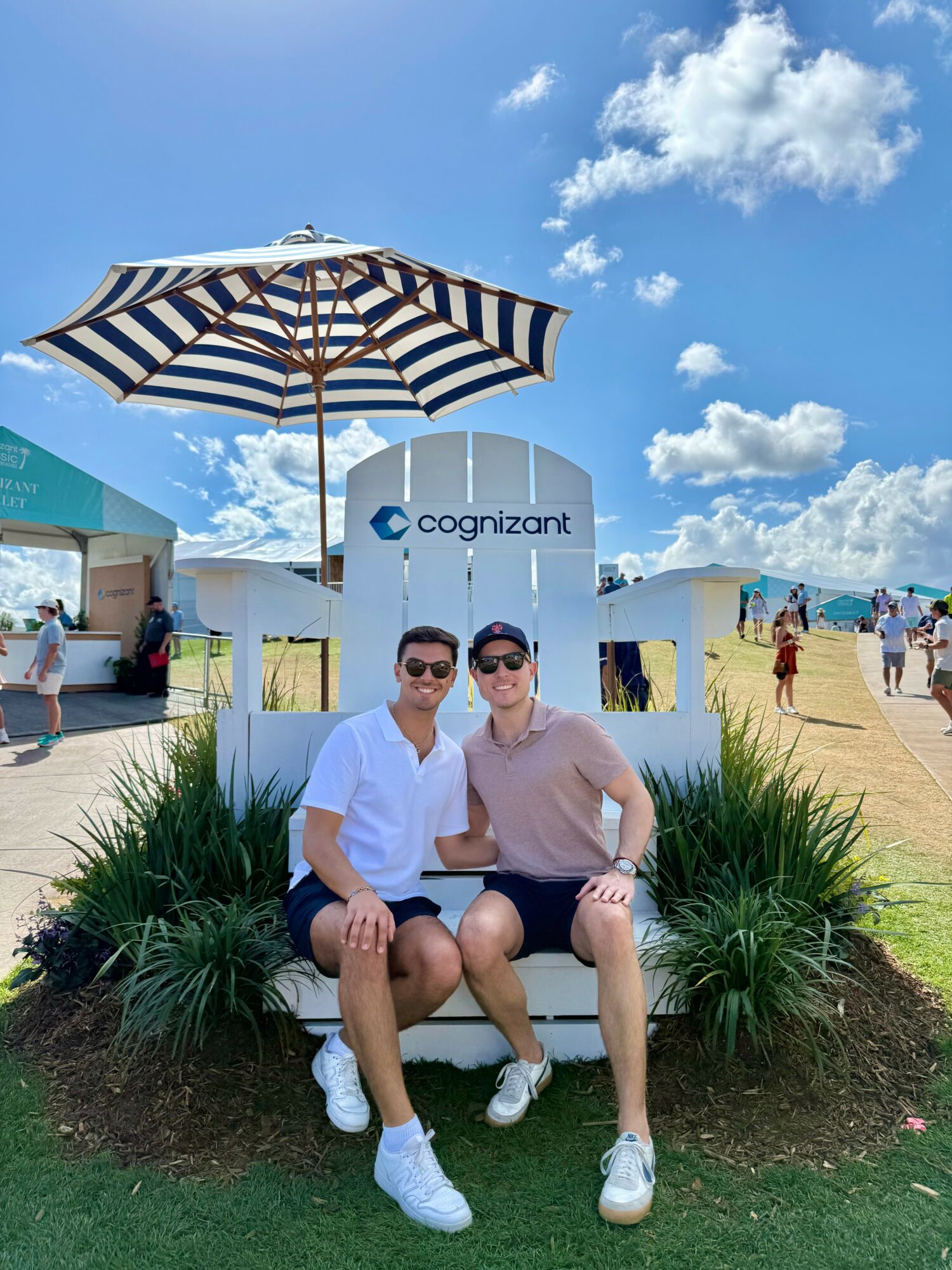 Two people sitting on a bench under a large striped umbrella at an outdoor event, with a blue sky and clouds in the background.