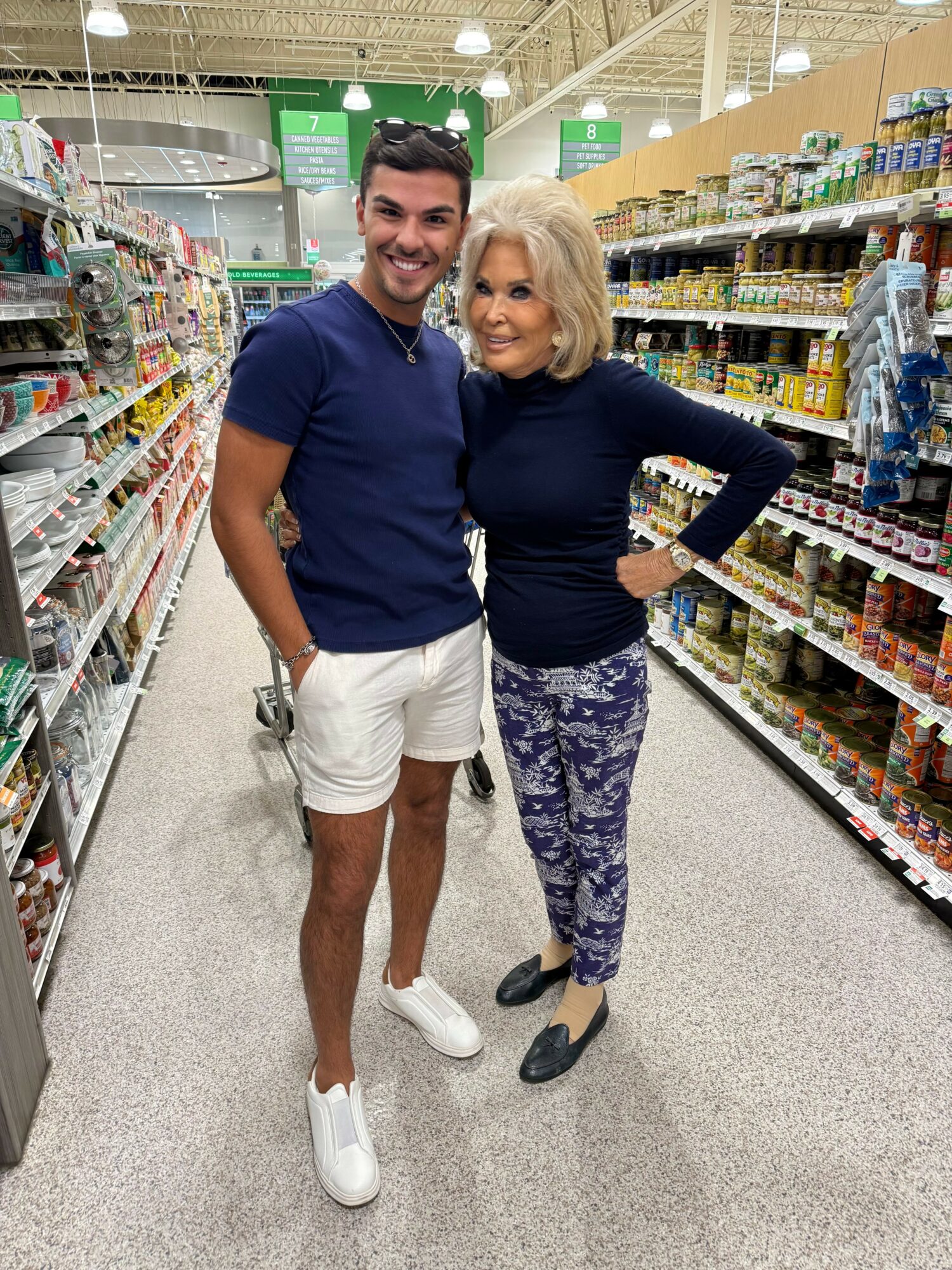 Two people smiling in a grocery store aisle, one with short dark hair and the other with shoulder-length blonde hair, both dressed casually.