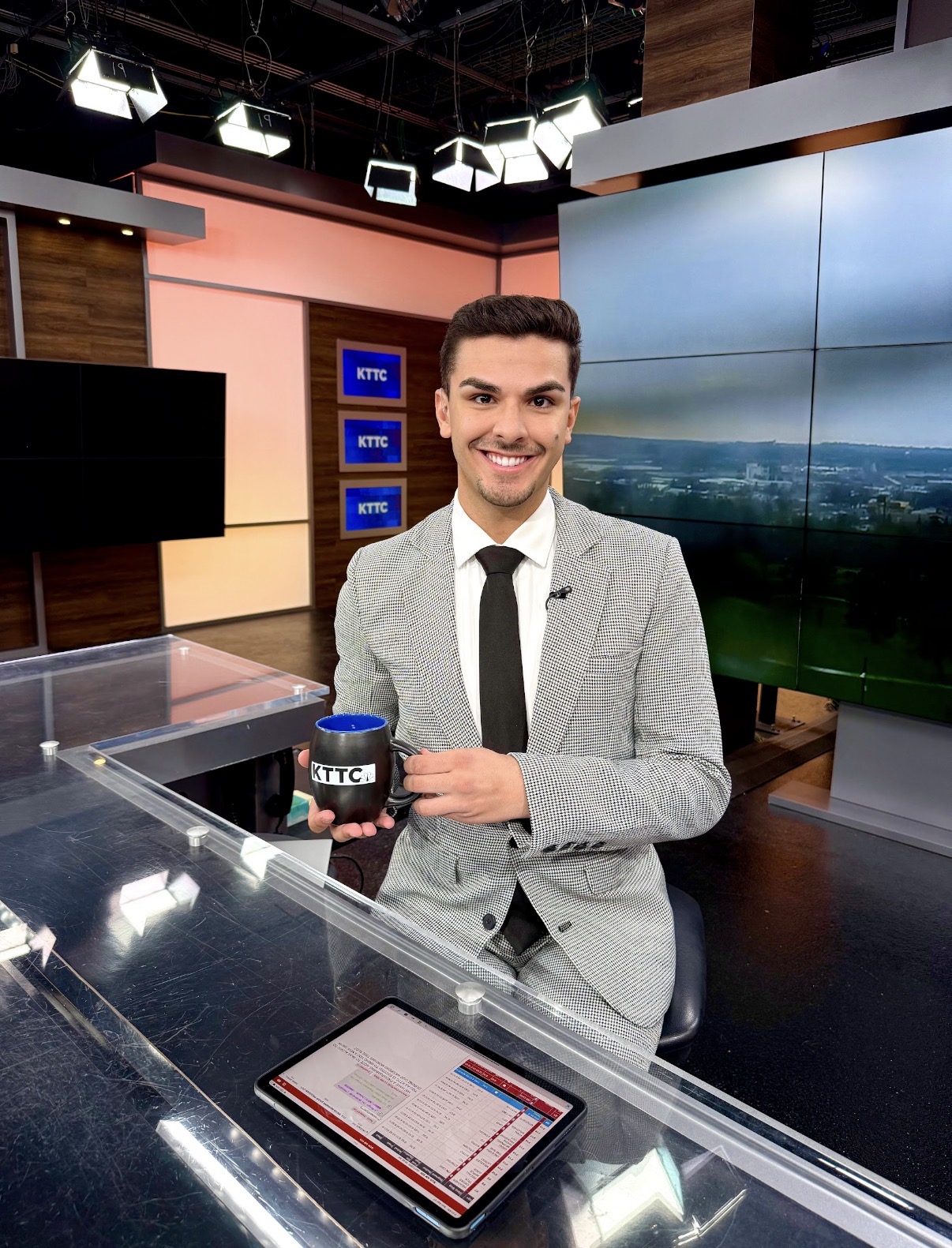 Man in a gray suit holding a coffee cup, smiling in a TV studio with screens and lighting, desk in foreground.