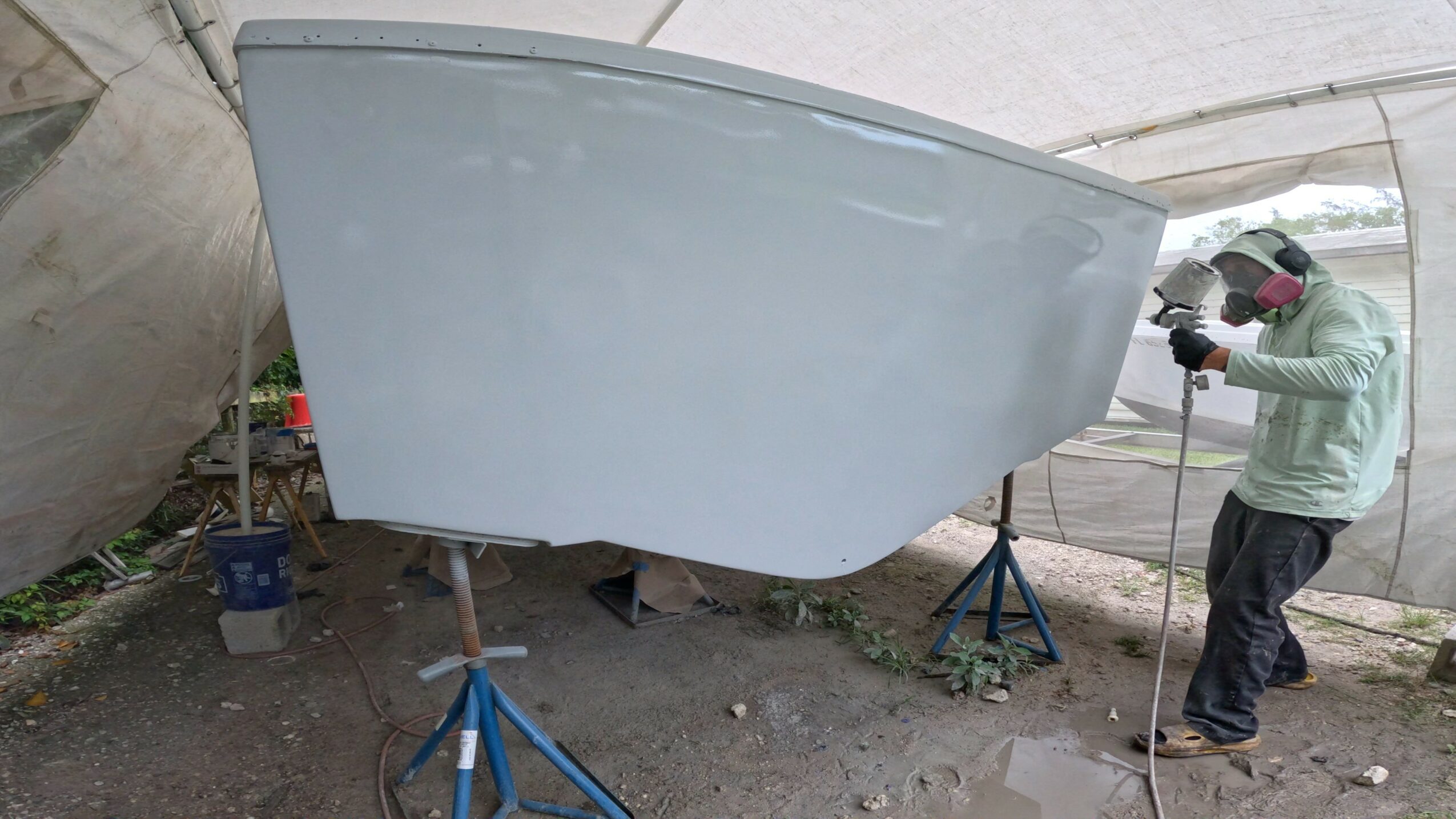 Person working on a boat inside a tent, with the boat on supports, wearing a jacket and gloves.