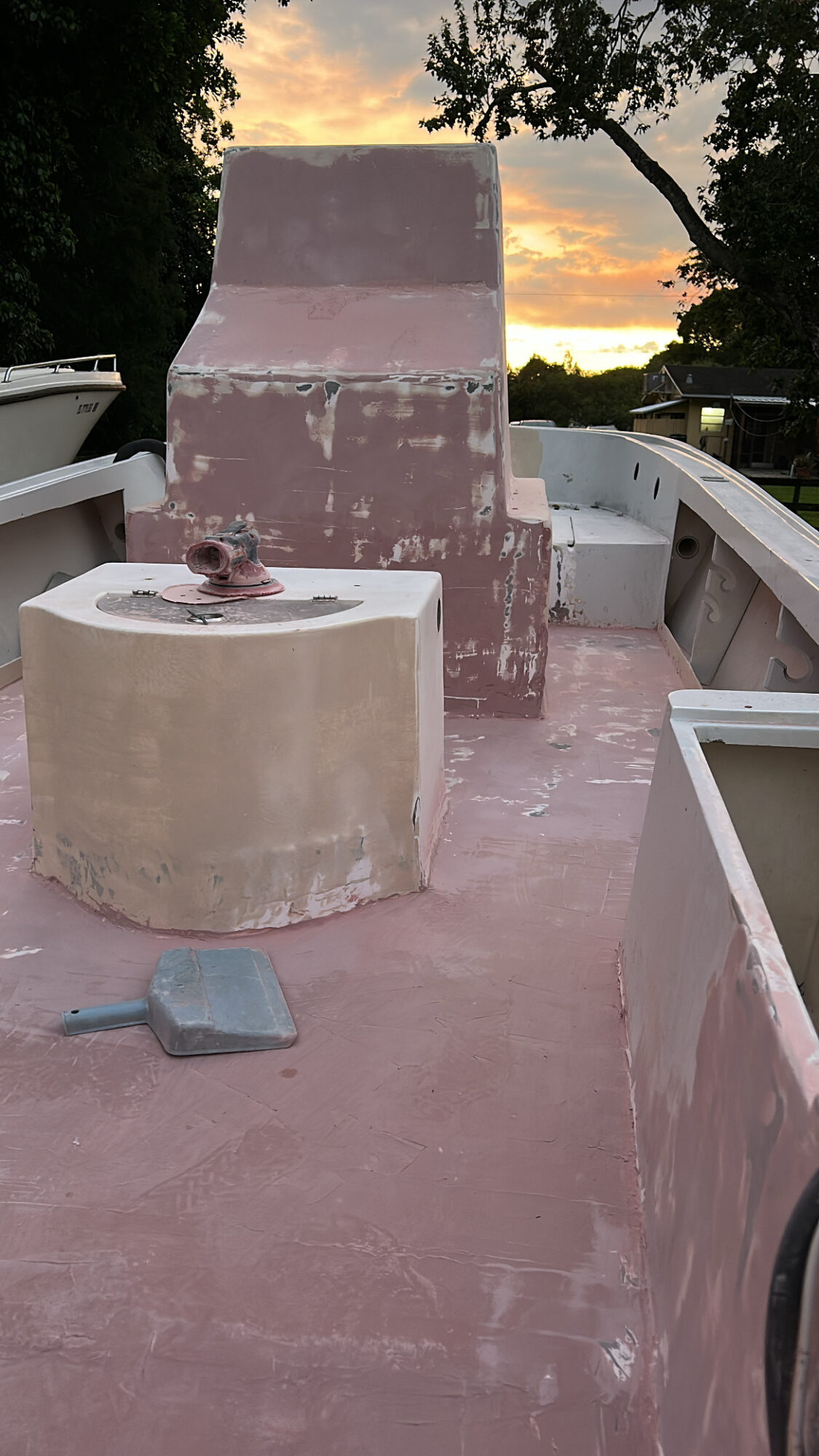 Boat deck with a large pink structure, a round beige object, and a small gray item, with trees and sunset in background.