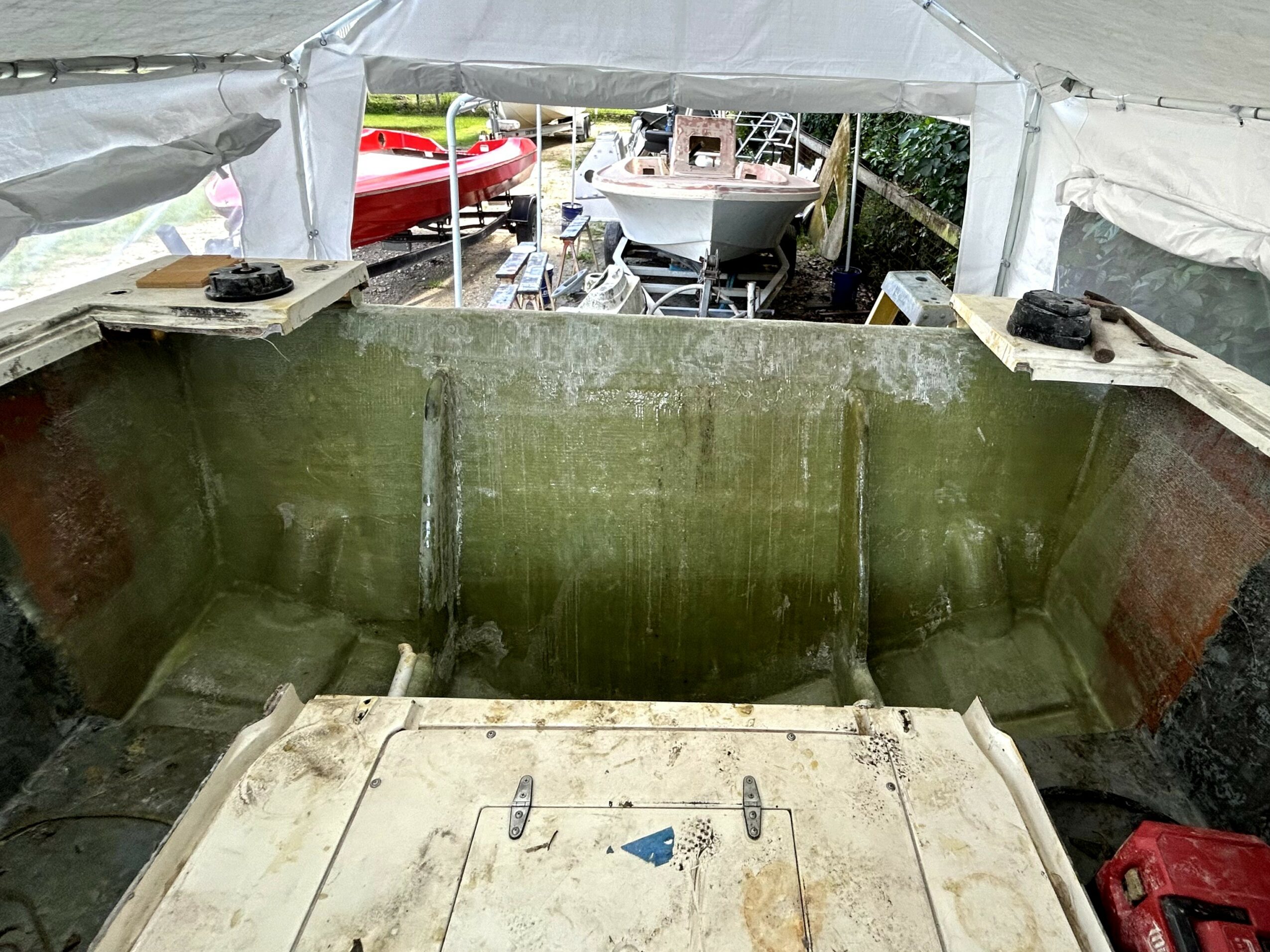 View from inside a boat under a tent, showing the boat's interior and a boatyard outside with boats and a red vehicle.