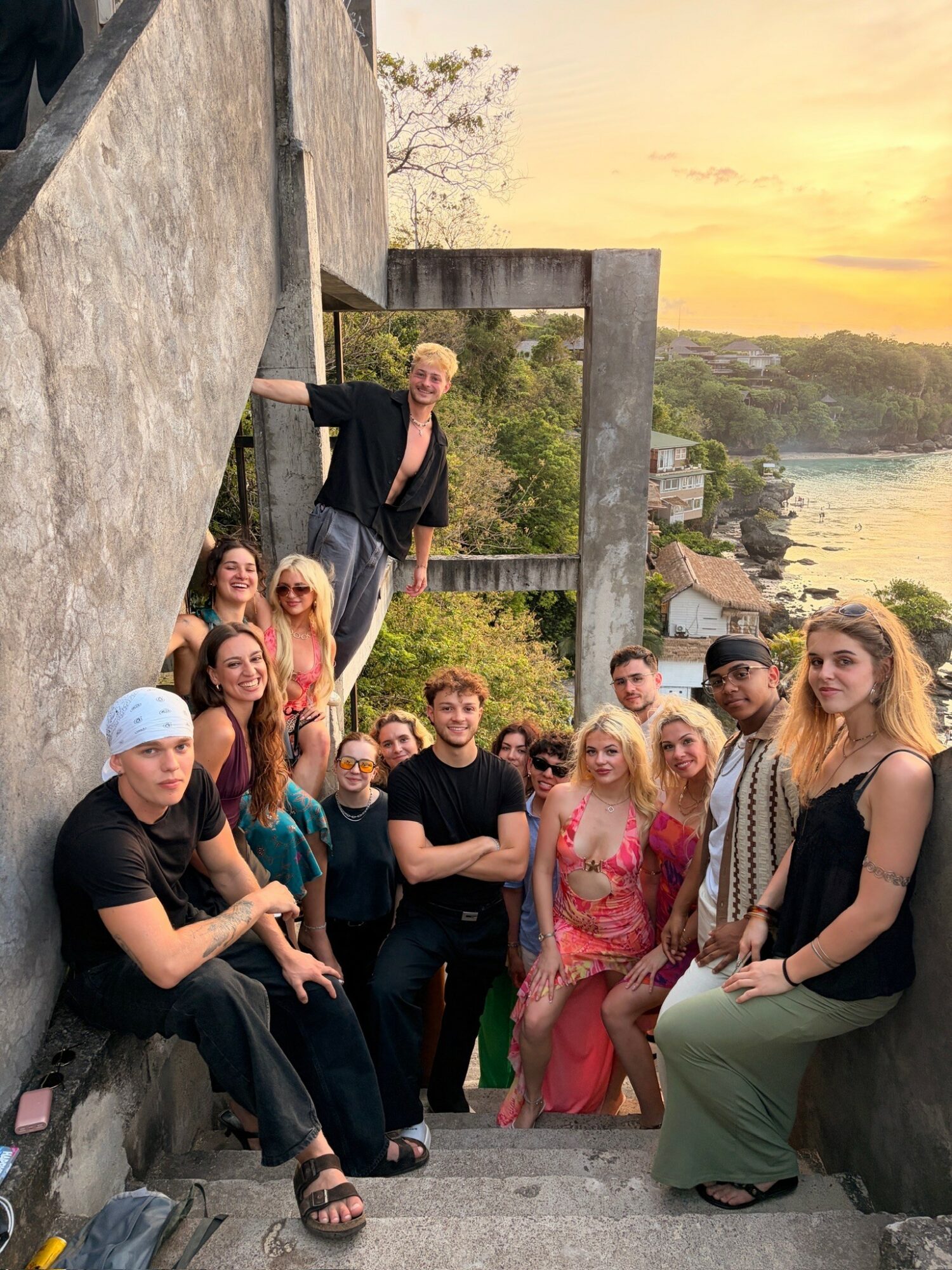Group of people posing on stairs outdoors with a body of water and sunset in background.