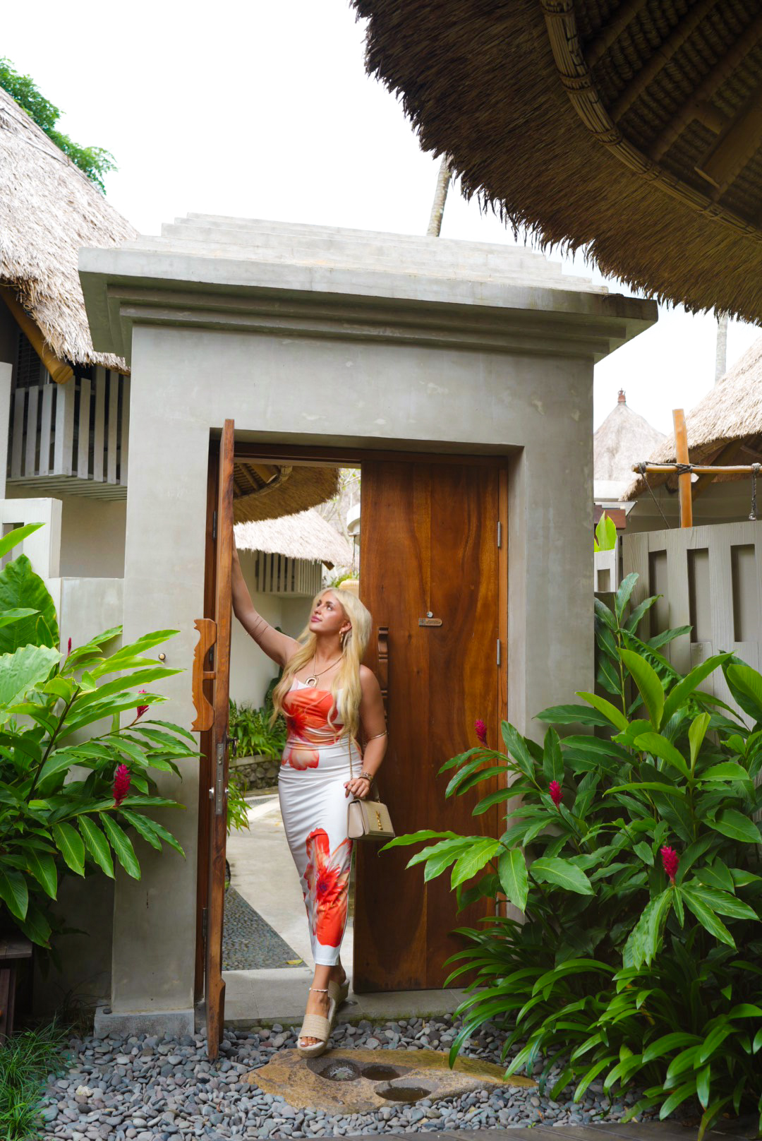 Woman in a colorful dress standing in a doorway surrounded by lush green plants, with a thatched roof visible at the top.