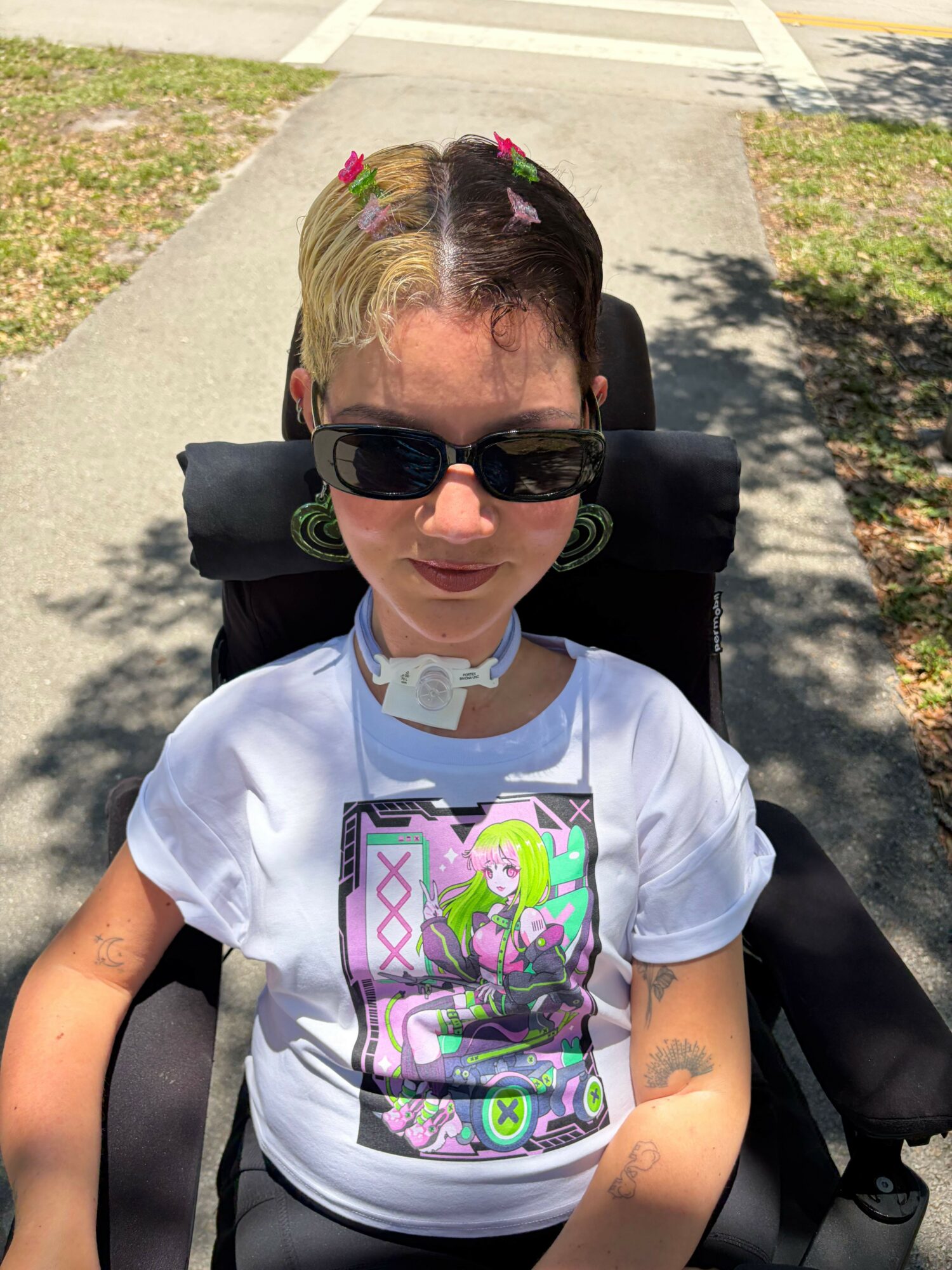 Person with short hair, sunglasses, and a white graphic t-shirt sitting outdoors on a sunny day.