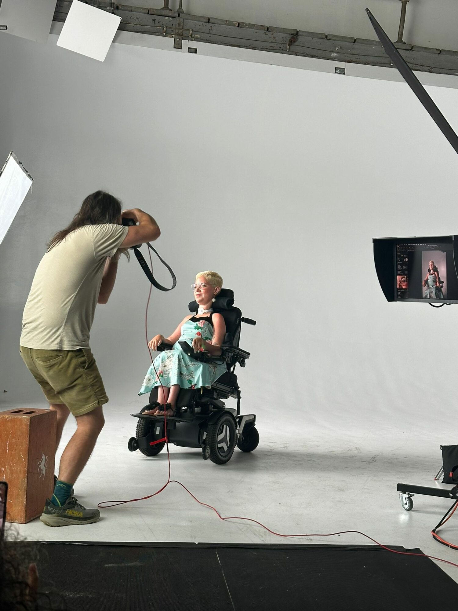Photographer taking picture of a young girl in a wheelchair against a white backdrop, studio setting, camera on tripod, cables on floor.