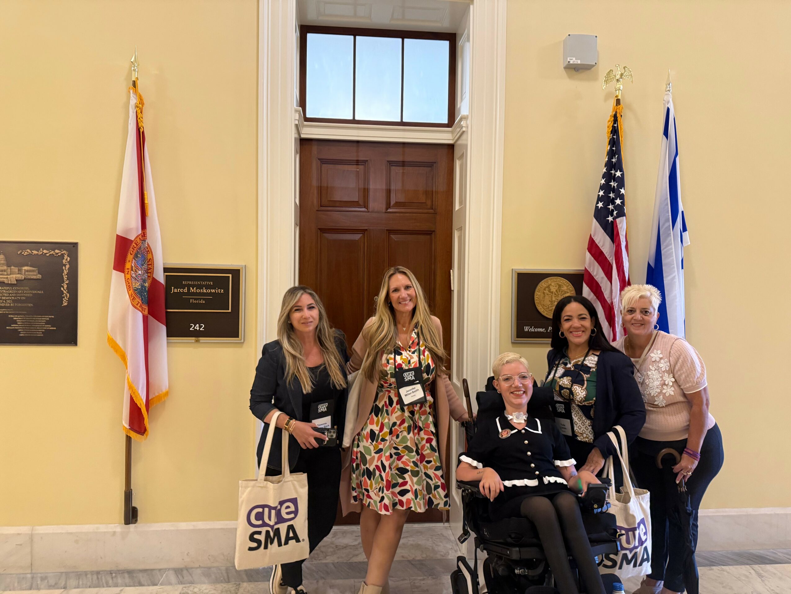 Four women and one person in a wheelchair pose in front of a door with flags on either side, inside a yellow-walled room.