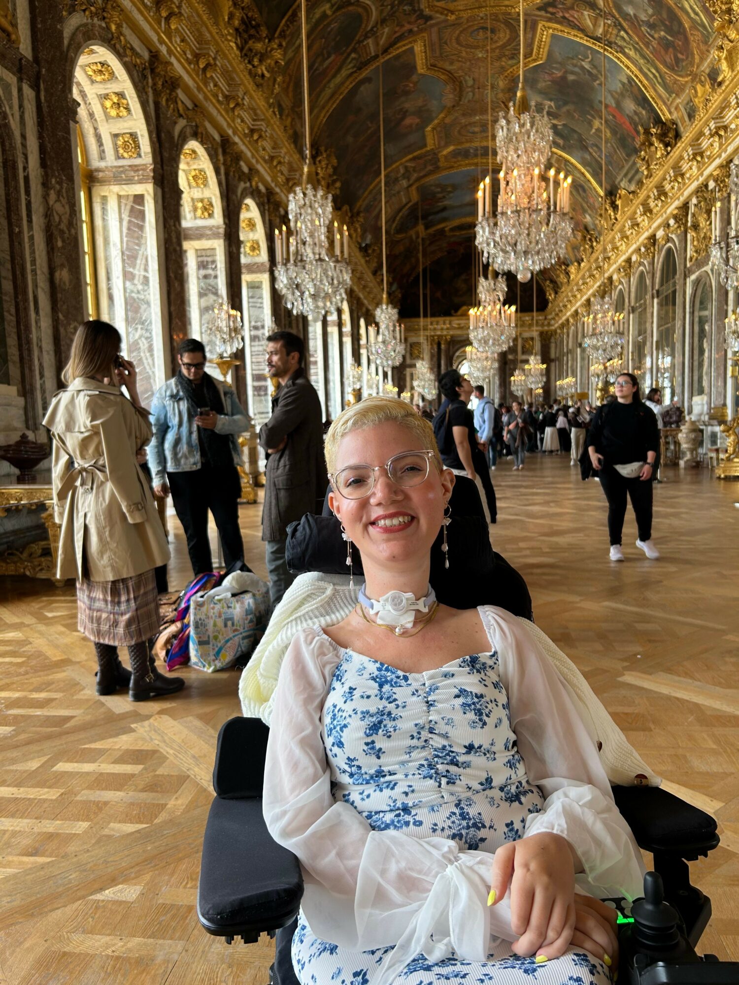 Woman sitting in a grand, ornate hall with chandeliers and gold decorations, smiling at the camera.