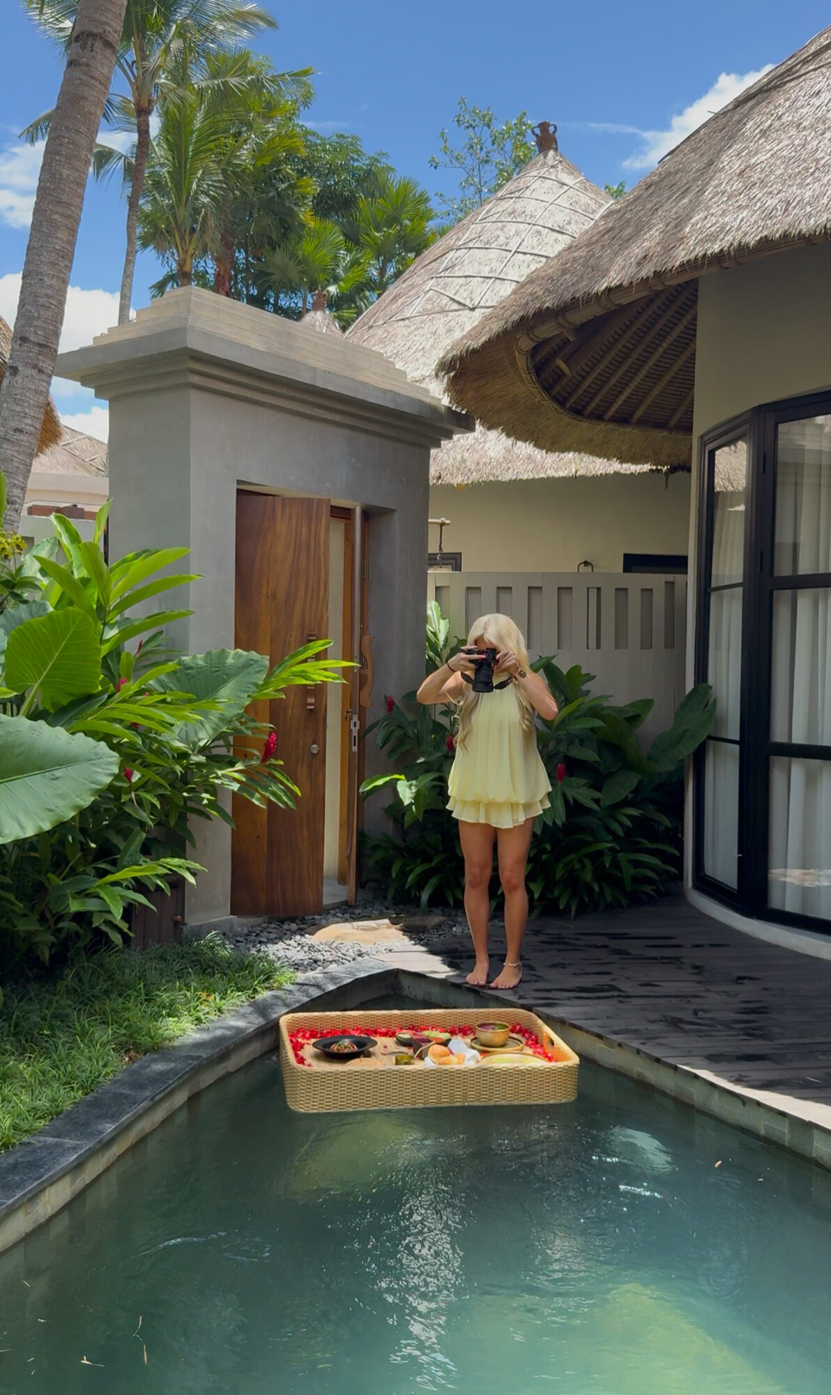 Person taking a photo near a pool with tropical plants and a thatched roof structure in the background.