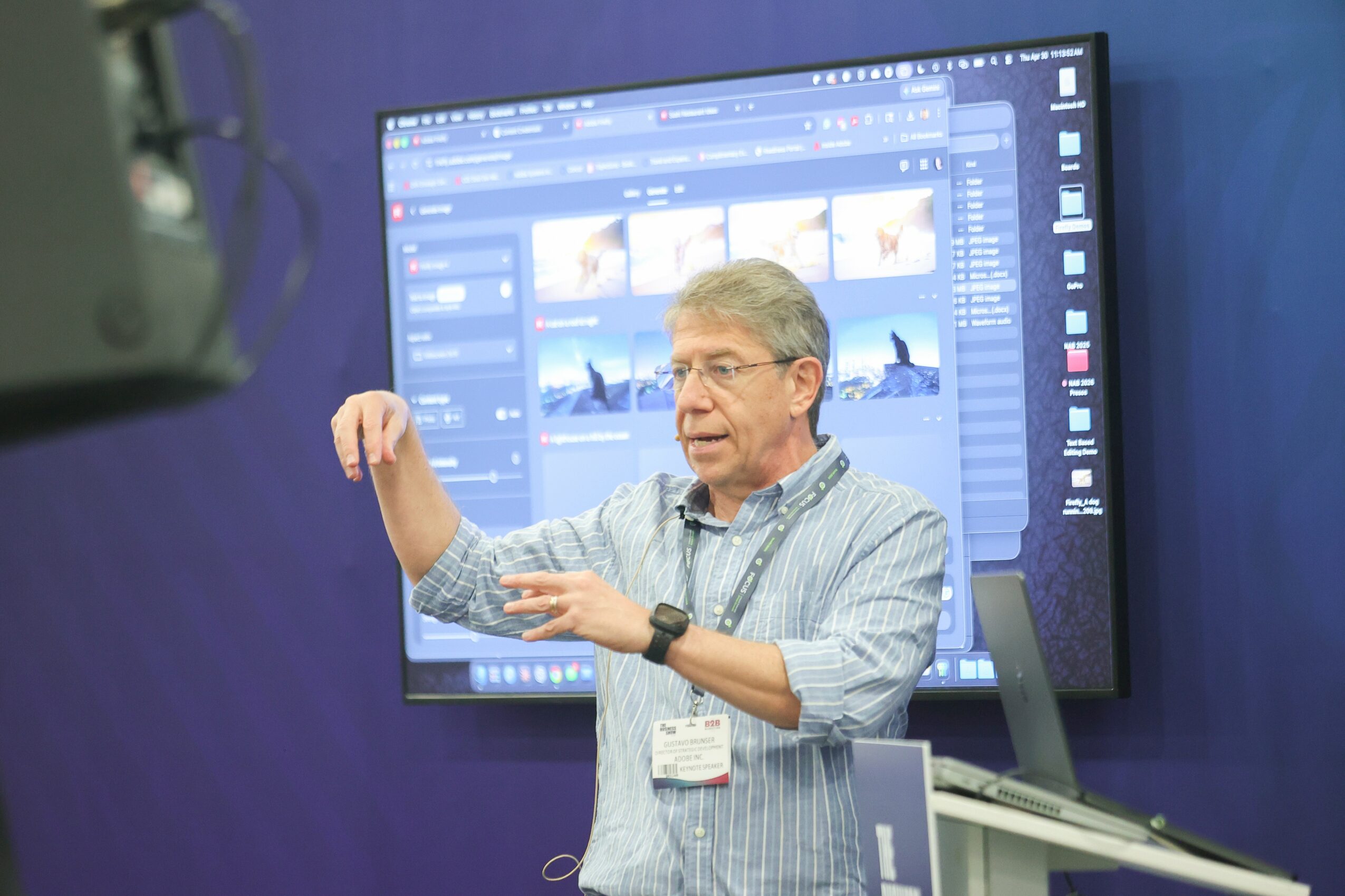 Older man with glasses and gray hair presenting in front of a large screen displaying photo editing software, wearing a striped shirt and a badge.