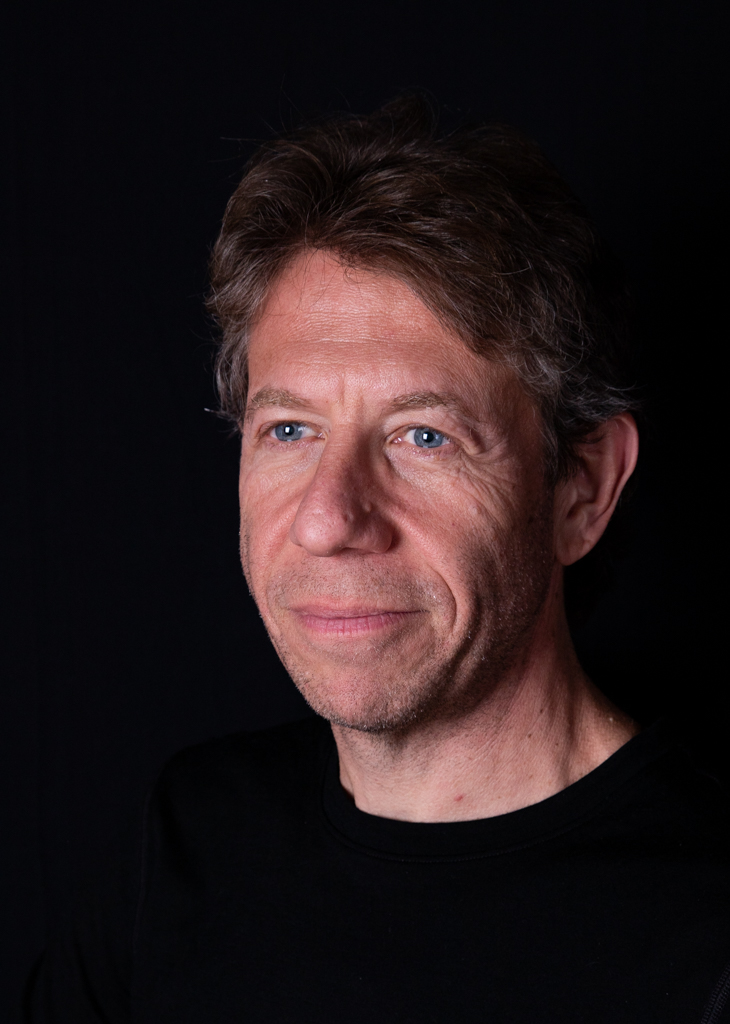 Portrait of a middle-aged man with light skin, blue eyes, and brown hair, wearing a black shirt, against a dark background.