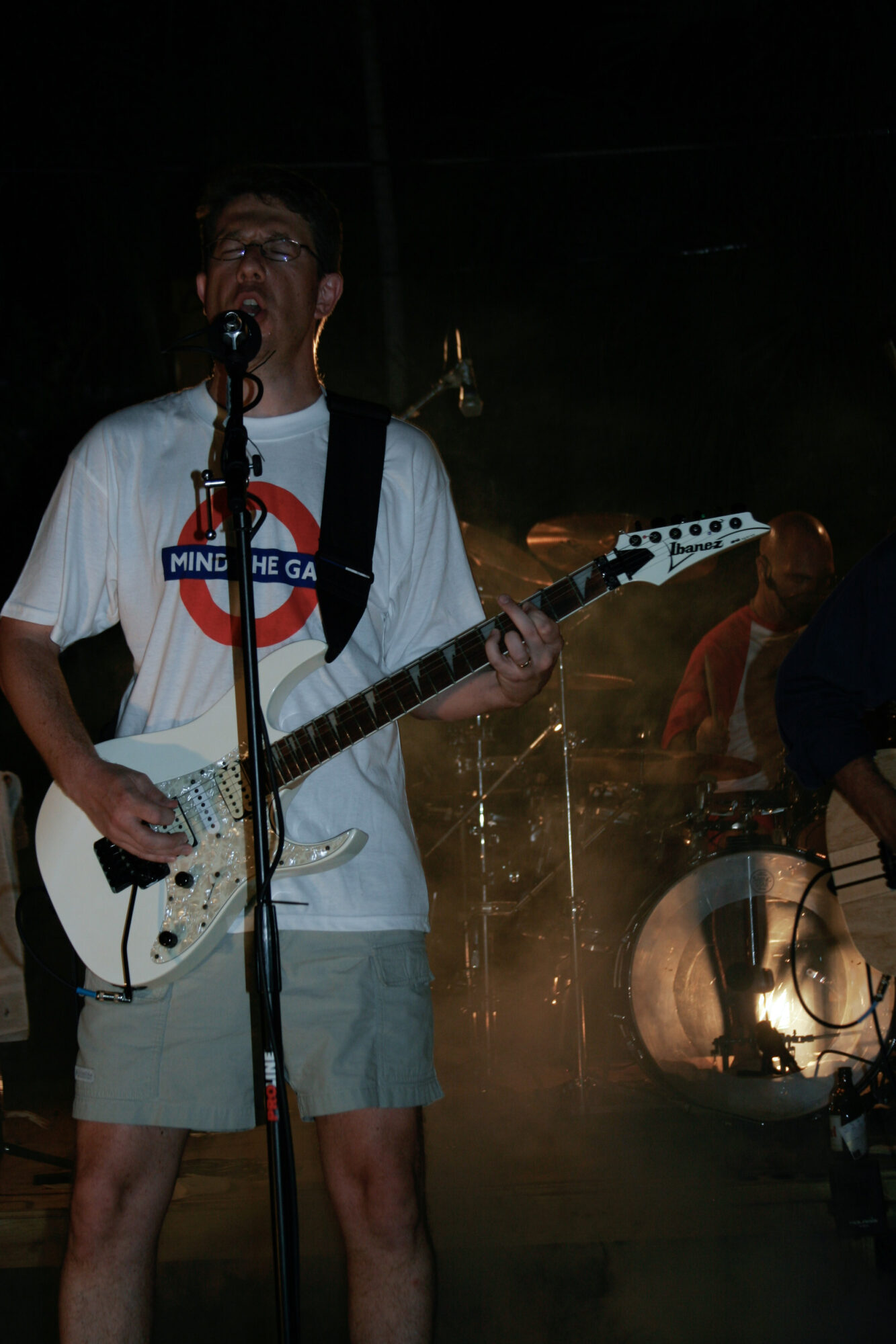 Person playing electric guitar and singing into microphone on stage with band members in background, dim lighting, and stage equipment.