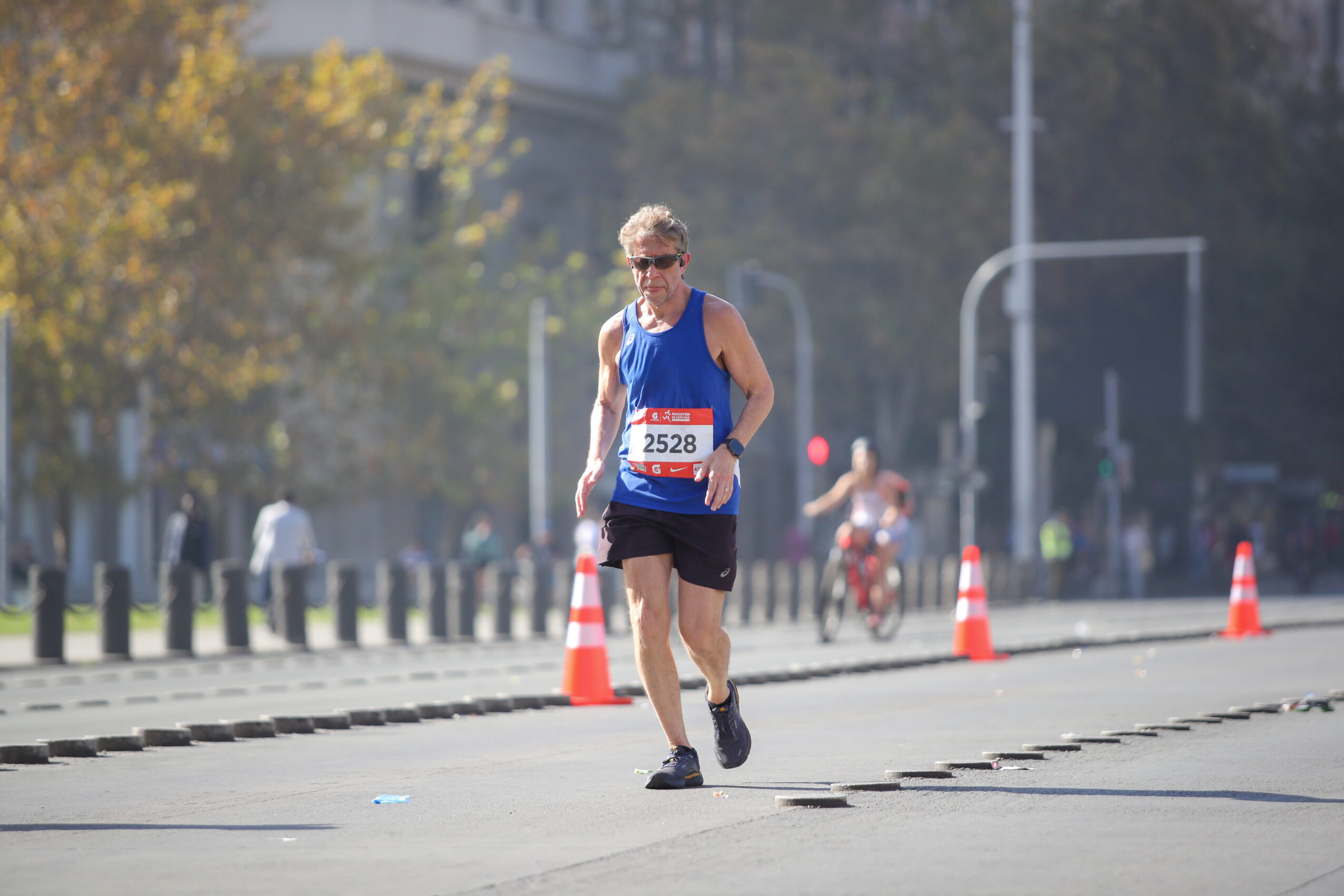A person running in a race, wearing a blue tank top, black shorts, sunglasses, and a race bib, on a city street.