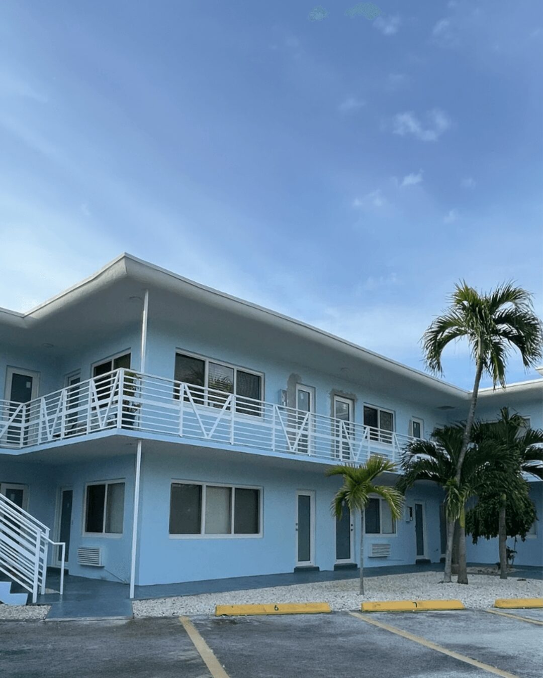 Two-story light blue building with white railings, surrounded by palm trees, under a blue sky with some clouds.
