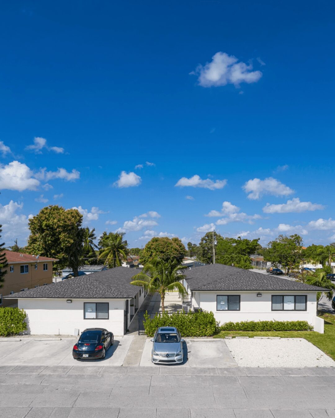 Two single-story houses with driveways and parked cars, trees, and a blue sky with clouds.