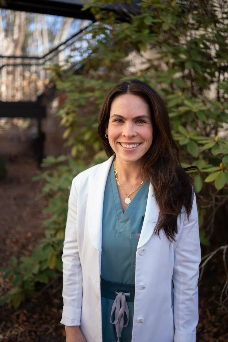 Woman with long dark hair smiling outdoors, wearing a white coat and blue scrubs, surrounded by green foliage.