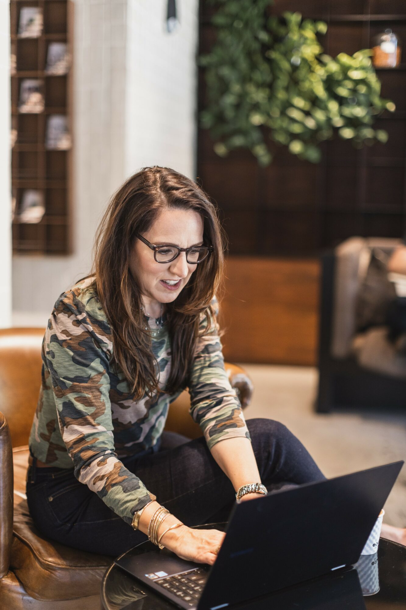 Woman with glasses using a laptop indoors, smiling, with a bookshelf and plant in background.