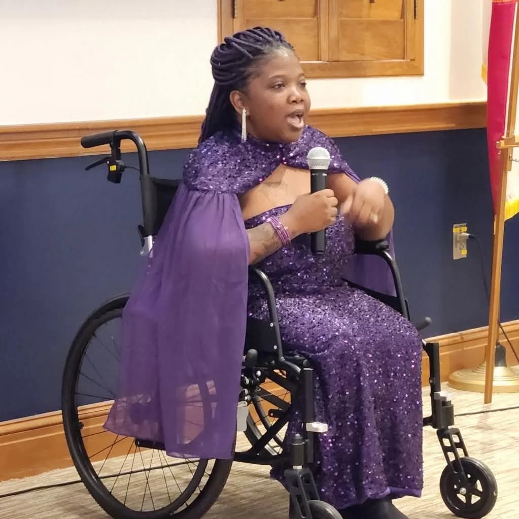 Woman in a wheelchair speaking into a microphone, wearing a purple dress and cape, indoors with wooden paneling.